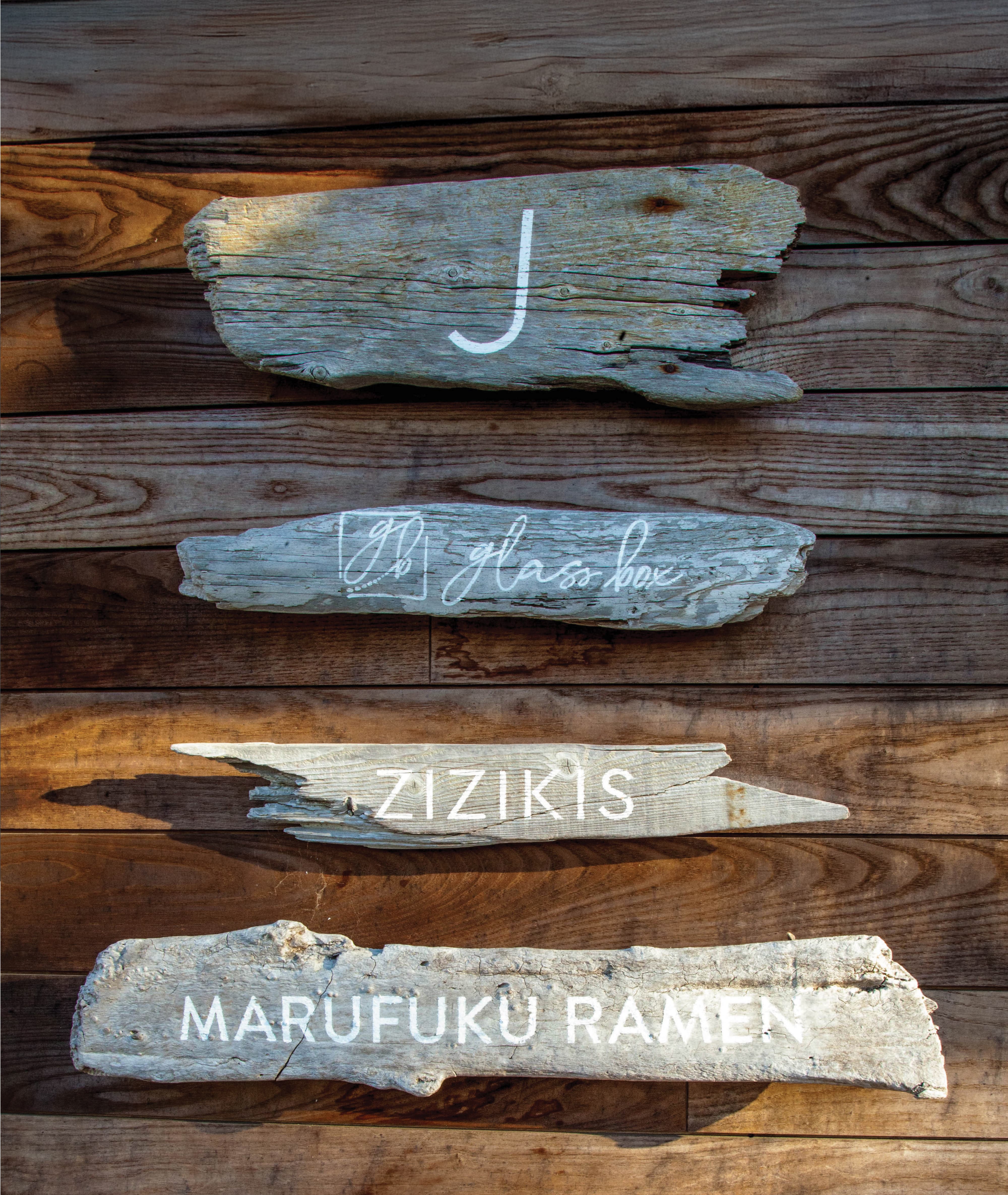 Four naturally weathered driftwood pieces mounted on a reclaimed wood plank wall, each hand-lettered in white with individual tenant identities — "J," Glass Box, Zizikis, and Marufuku Ramen — serving as organic, material-driven wayfinding and tenant identification signs that exemplify RSM Design's specialty graphics, placemaking, and experiential graphics and branded environments approach within the food and beverage and mixed-use markets