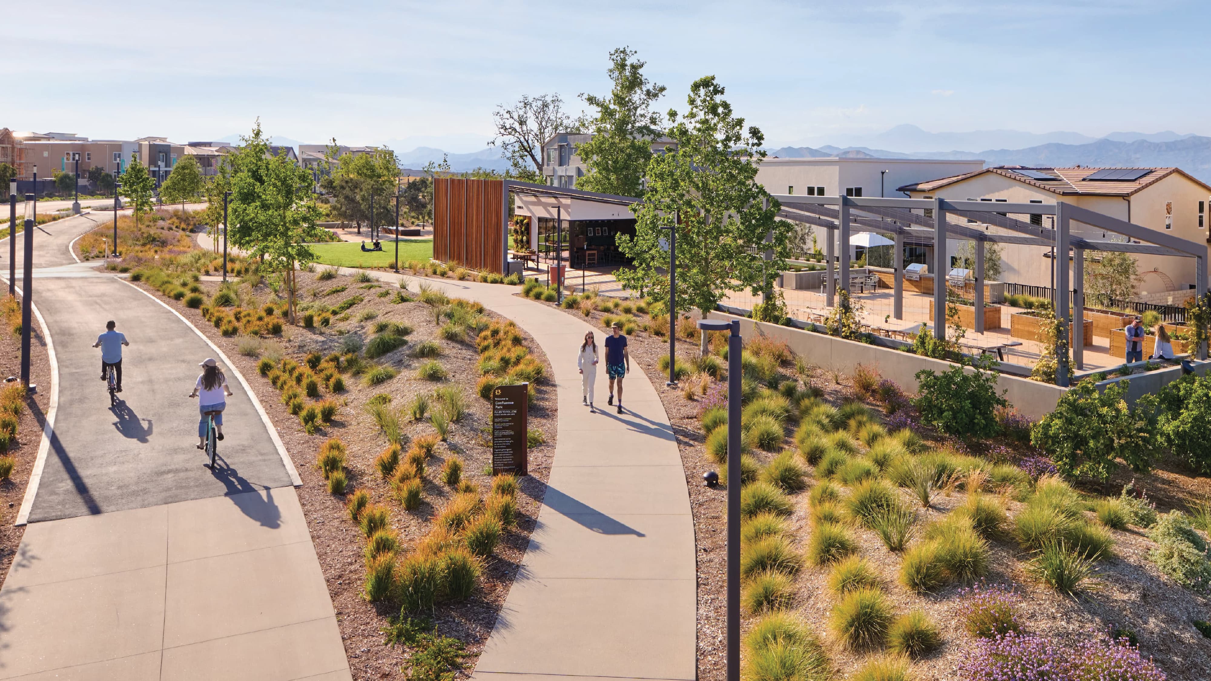 Elevated view of Confluence Park with a curved bike path and walking path through native grasses, people biking and walking, a wayfinding sign, and a community pavilion and shaded outdoor lounge area with residential buildings and mountains beyond.