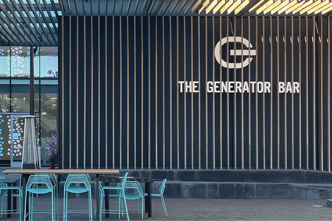The Generator Bar exterior facade with dimensional logo signage on wood slat and dark panel wall, outdoor patio seating, and decorative glass graphic panels at San Carlos by RSM Design