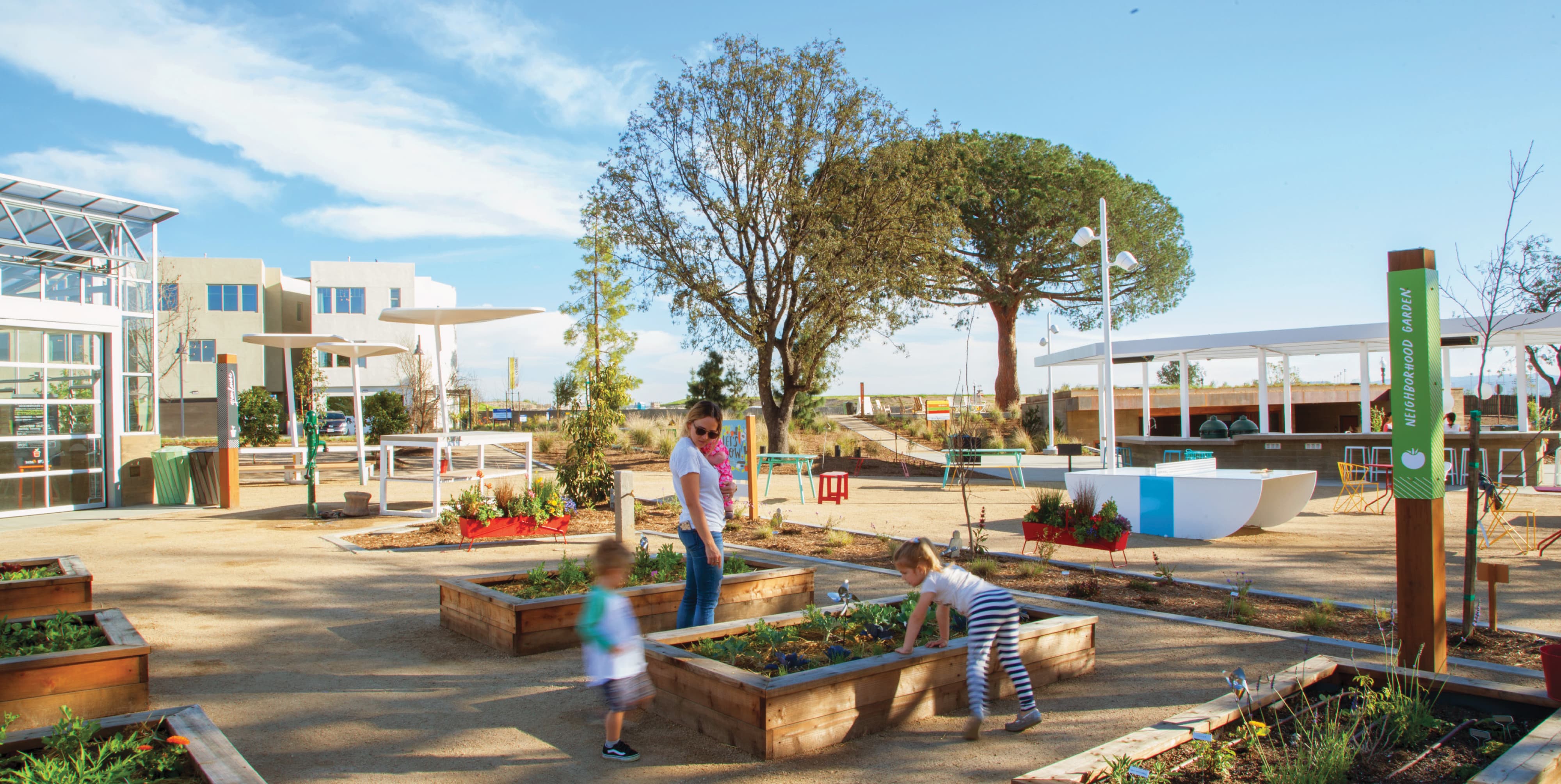 RSM Design placemaking, wayfinding signage, and experiential graphics for FivePoint at Great Park Neighborhoods in Irvine, California, featuring a wide view of an activated community commons with a Neighborhood Garden identification post sign, raised wood planting beds with a mother and children exploring, colorful outdoor furniture, shade structures, a community pavilion, and a mature tree canopy against a blue sky, demonstrating how environmental graphic design, specialty graphics, and placemaking work together to create meaningful, human-centered outdoor gathering spaces and community identity for residential community and park markets.