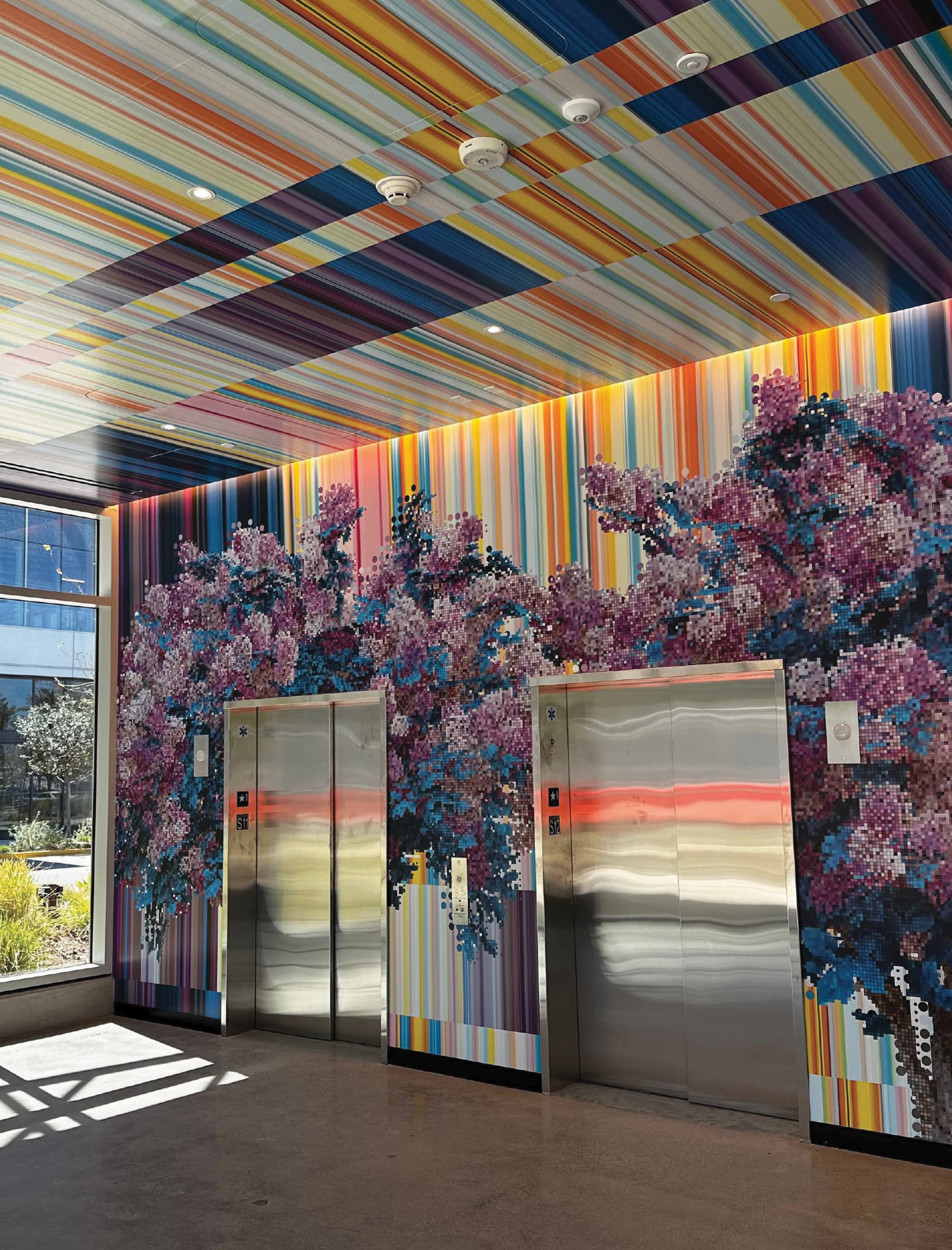 Elevator lobby with two stainless steel elevator doors set beneath a colorful striped ceiling and wall mural of pixelated purple and blue floral blossoms transitioning into a rainbow-striped pattern.