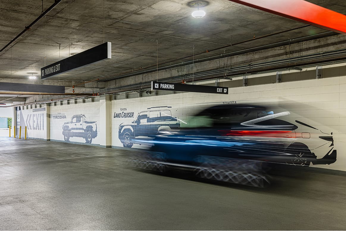 Interior parking garage wayfinding signage and Toyota-branded vehicle murals featuring Tacoma and Land Cruiser graphics lining the drive aisle walls at Toyota Music Factory in Irving, Texas — RSM Design wayfinding and signage design, experiential graphics, branded environments, and master sign programs for parking garage and mixed-use destinations.
