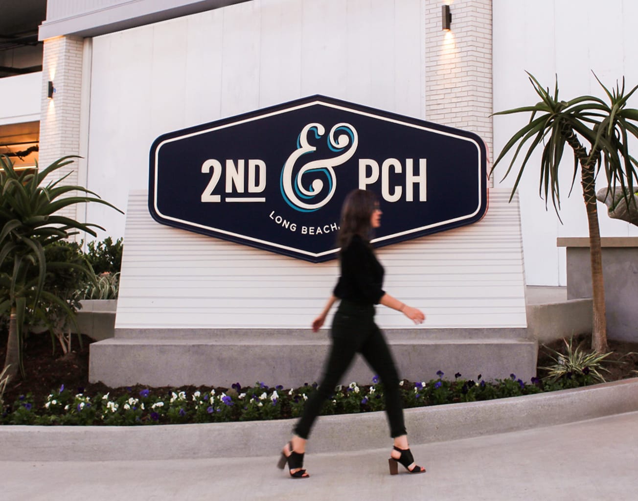 Pedestrian walking past a large-scale dimensional wall-mounted monument sign for 2nd & PCH in Long Beach, California, featuring the navy coffin-shaped logo cabinet with cream lettering and a decorative teal ampersand set within a landscaped planter — showcasing RSM Design's environmental graphic design, placemaking, and branded environments for a coastal waterfront retail and mixed-use destination.
