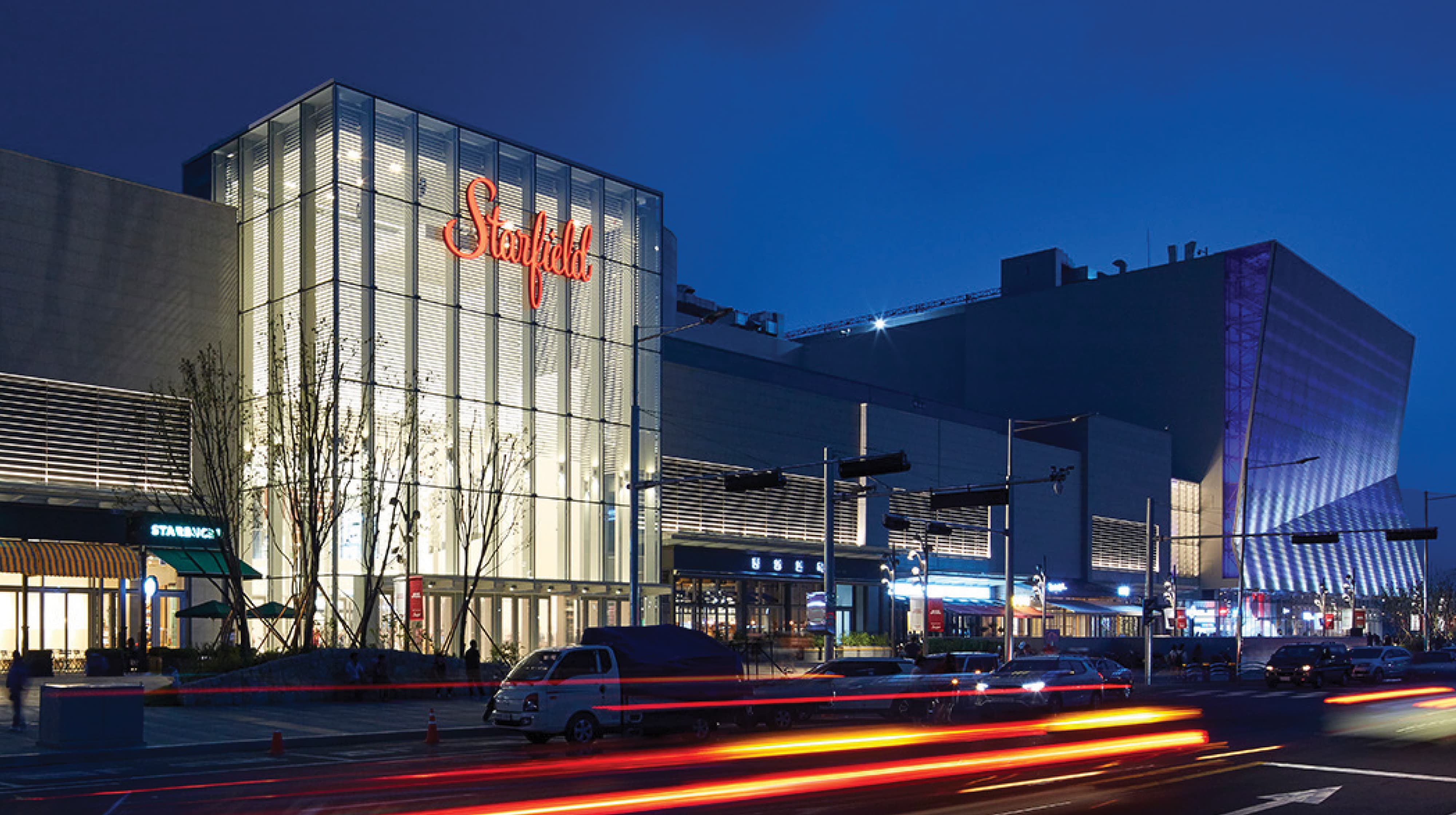 Nighttime exterior of the Starfield mall with an illuminated glass tower displaying the red 'Starfield' script logo, storefronts including Starbucks, and light trails from passing traffic in the foreground.