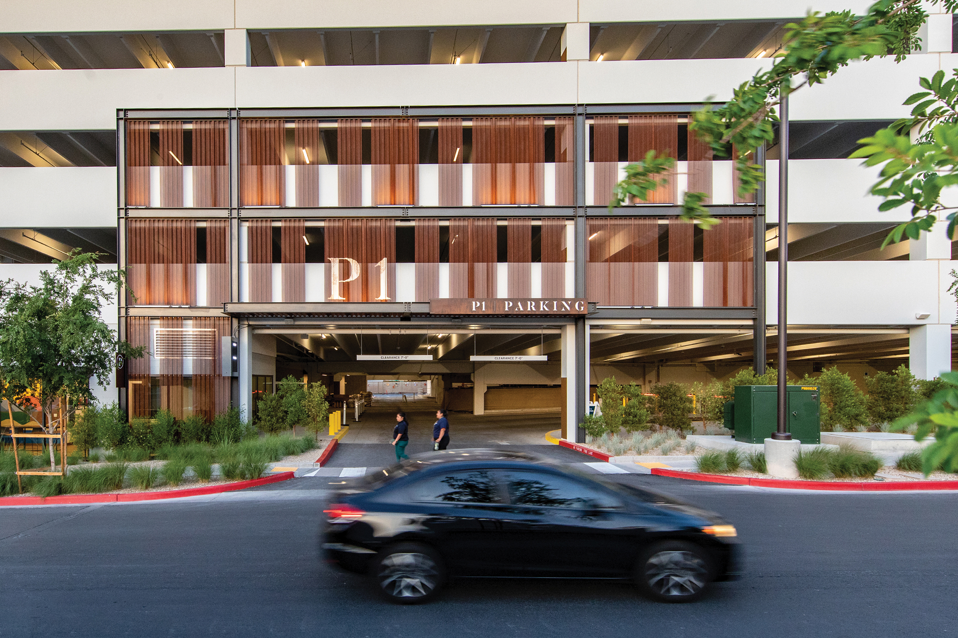 Exterior view of the P1 parking structure at Uncommons, with a car passing in the foreground. The facade features warm copper-toned vertical screen panels with a large illuminated "P1" sign. Two attendants stand near the entrance below a "P1 Parking" header sign. Ornamental grasses and shrubs line the drive aisle, with a multi-level open parking deck visible above.