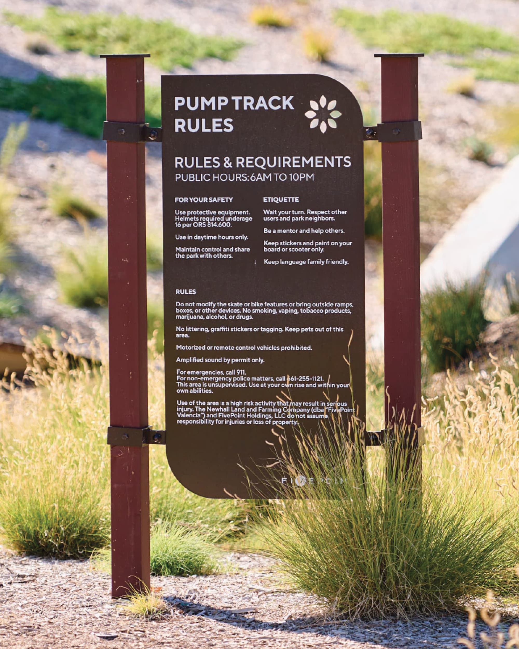 Pump Track Rules sign listing safety guidelines, etiquette, and requirements, mounted between two dark red posts amid tall grasses and desert landscaping.