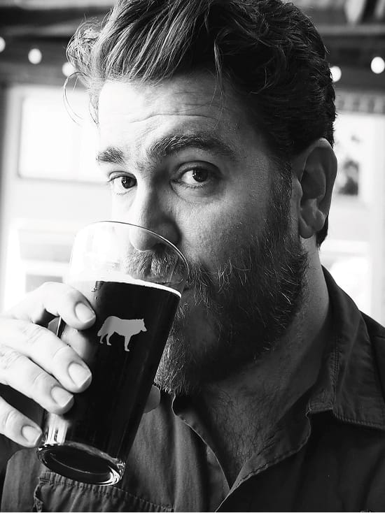 Black and white portrait of a bearded man drinking a dark craft beer from a pint glass with a bear logo, candid close-up lifestyle photography in a bar setting.