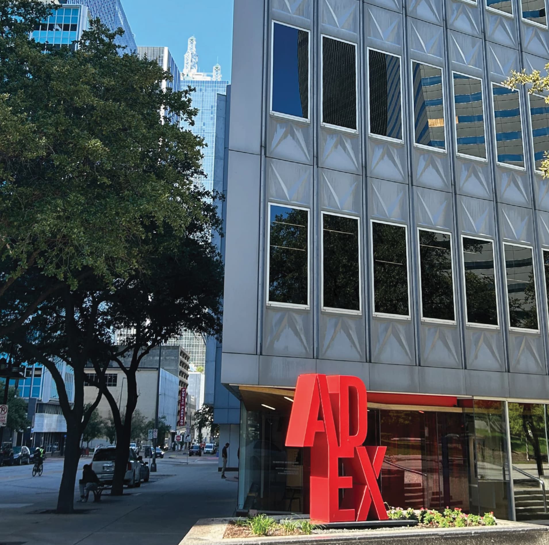 RSM Design landmark identity sculpture and exterior signage for the Architecture and Design Exchange in downtown Dallas, featuring the bold red freestanding dimensional ADEX letter forms positioned at street level against the landmark modernist glass curtain wall building facade, with a mature tree-lined urban streetscape, cyclist, and downtown Dallas skyline visible in the background, demonstrating how environmental graphic design, public art, placemaking, and landmark identity signage transform a civic institution into a recognizable, human-centered destination within a cultural, civic, and urban streetscape context.