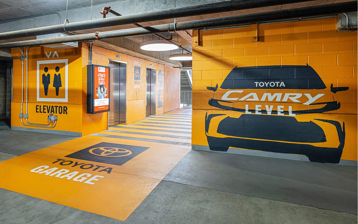 Toyota Camry branded parking garage elevator lobby with immersive orange wall murals, large-scale vehicle graphics, pictogram elevator signage, floor graphics, digital display, and Toyota Garage identity at Toyota Music Factory in Irving, Texas — RSM Design experiential graphics, wayfinding and signage design, digital integration, branded environments, and master sign programs for parking garage and mixed-use destinations.
