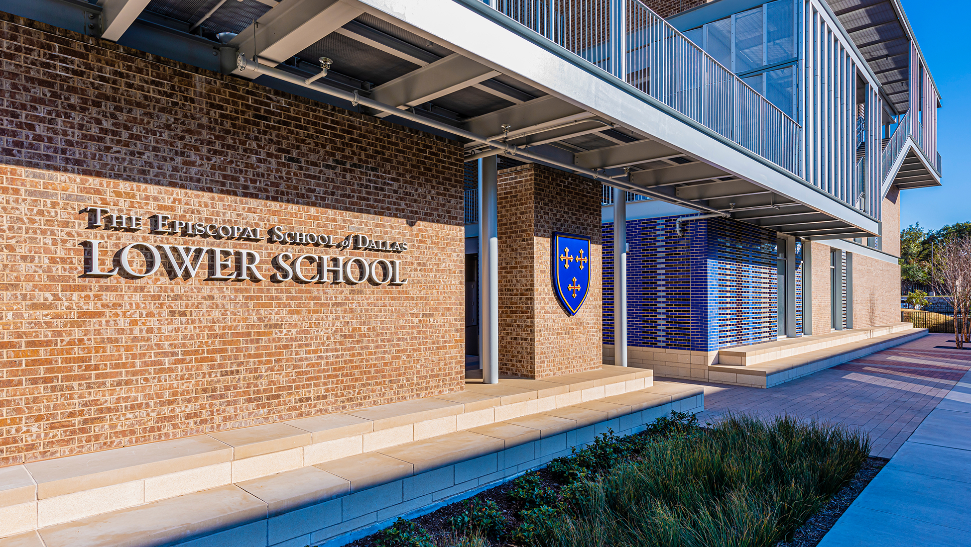 Brick exterior of 'The Episcopal School of Dallas, Lower School' with dimensional lettering and a blue crest shield bearing four gold crosses, beneath a covered walkway with a mosaic blue-brick accent wall and landscaped entrance.