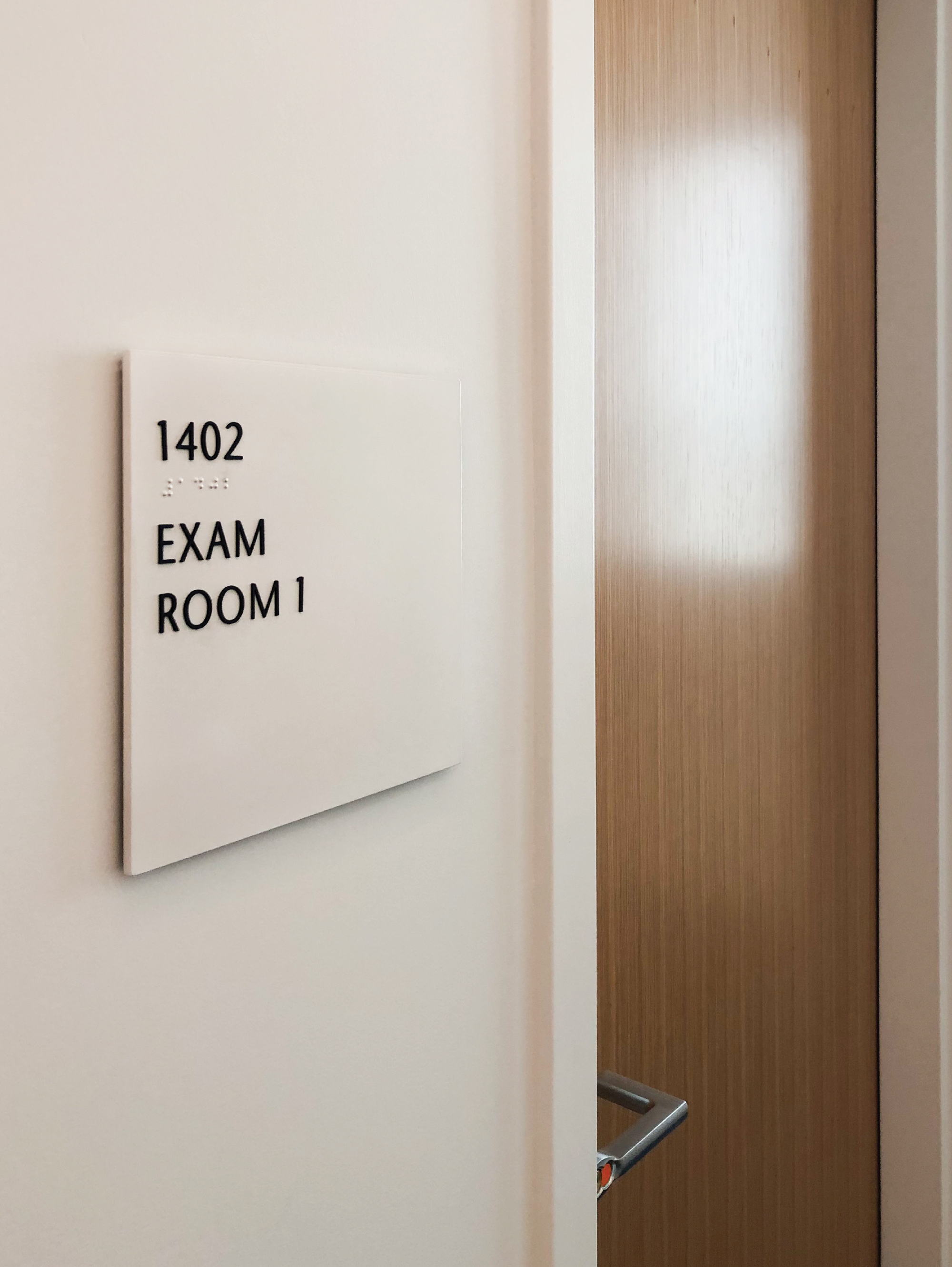 Accessible room identification signage with braille for Exam Room 1 in a higher education facility, illustrating environmental graphic design, ADA-compliant wayfinding, and integrated signage systems within the built environment.