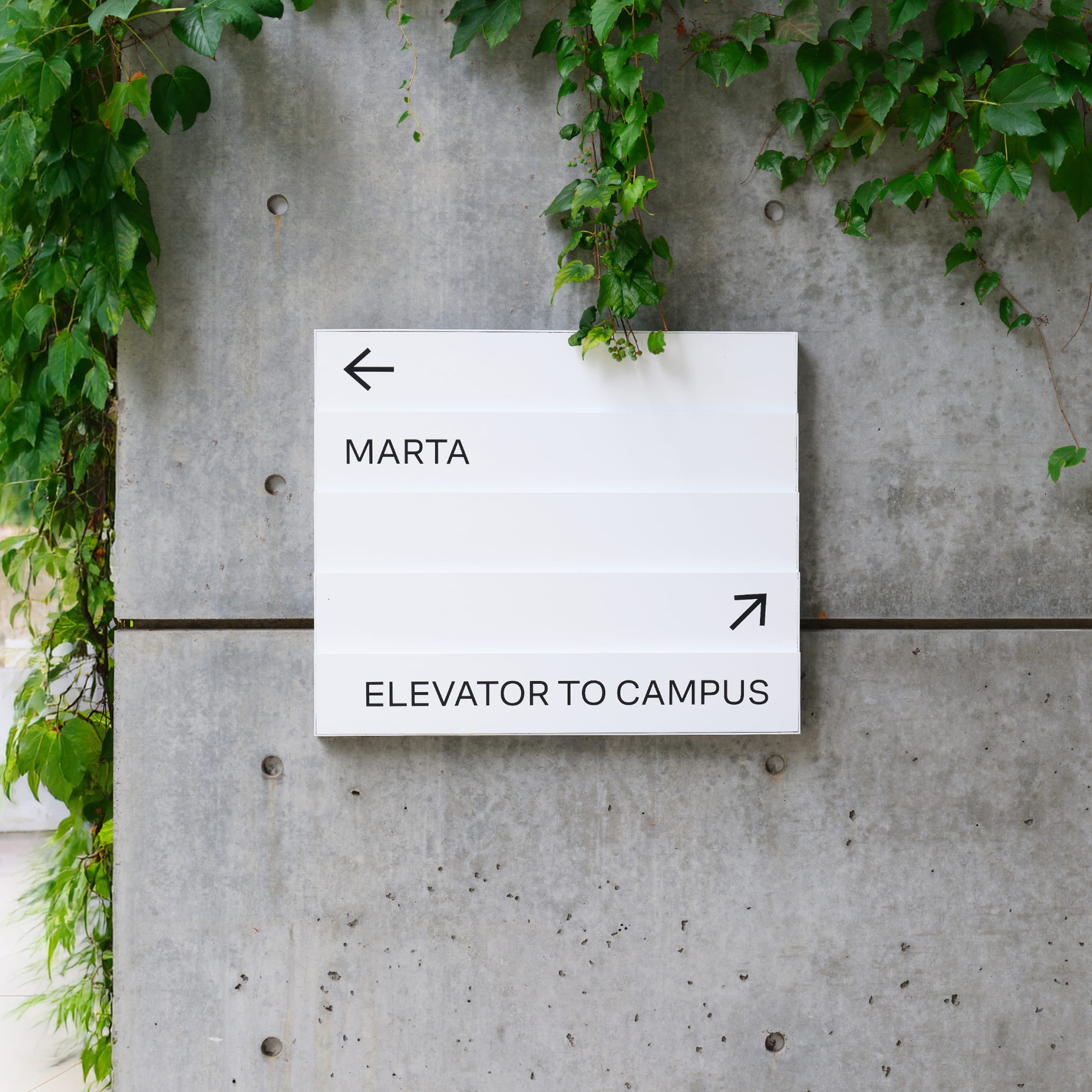 Wall-mounted directional sign with arrows pointing to MARTA transit and the elevator to campus, set against an ivy-covered concrete wall at the Woodruff Arts Center – campus wayfinding and environmental graphic design by RSM Design.