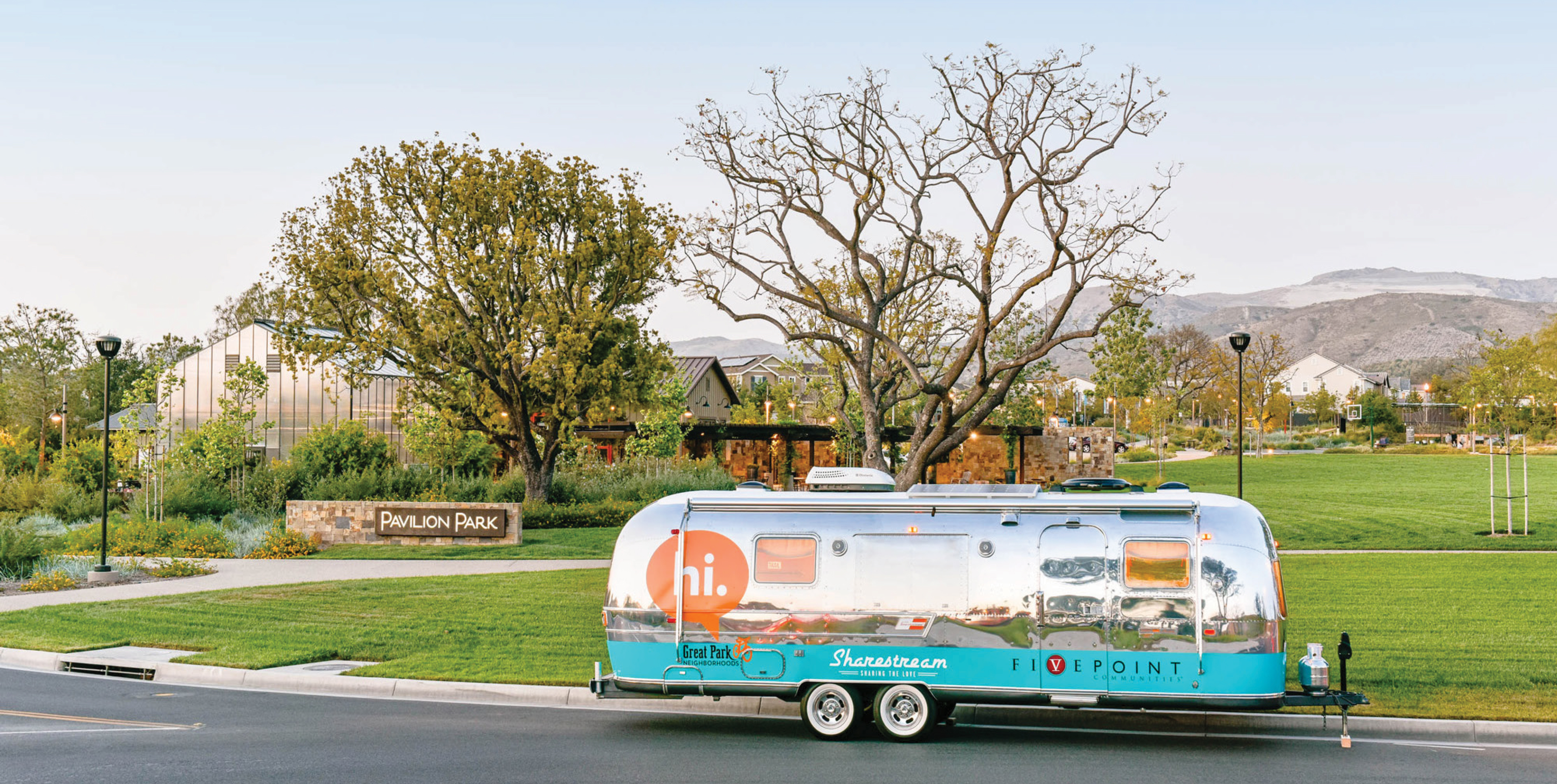 Silver Airstream trailer branded 'hi.' and 'Sharestream' by FivePoint parked on the street in front of the Pavilion Park sign and community greenspace, with a glass greenhouse building and mountains in the background.