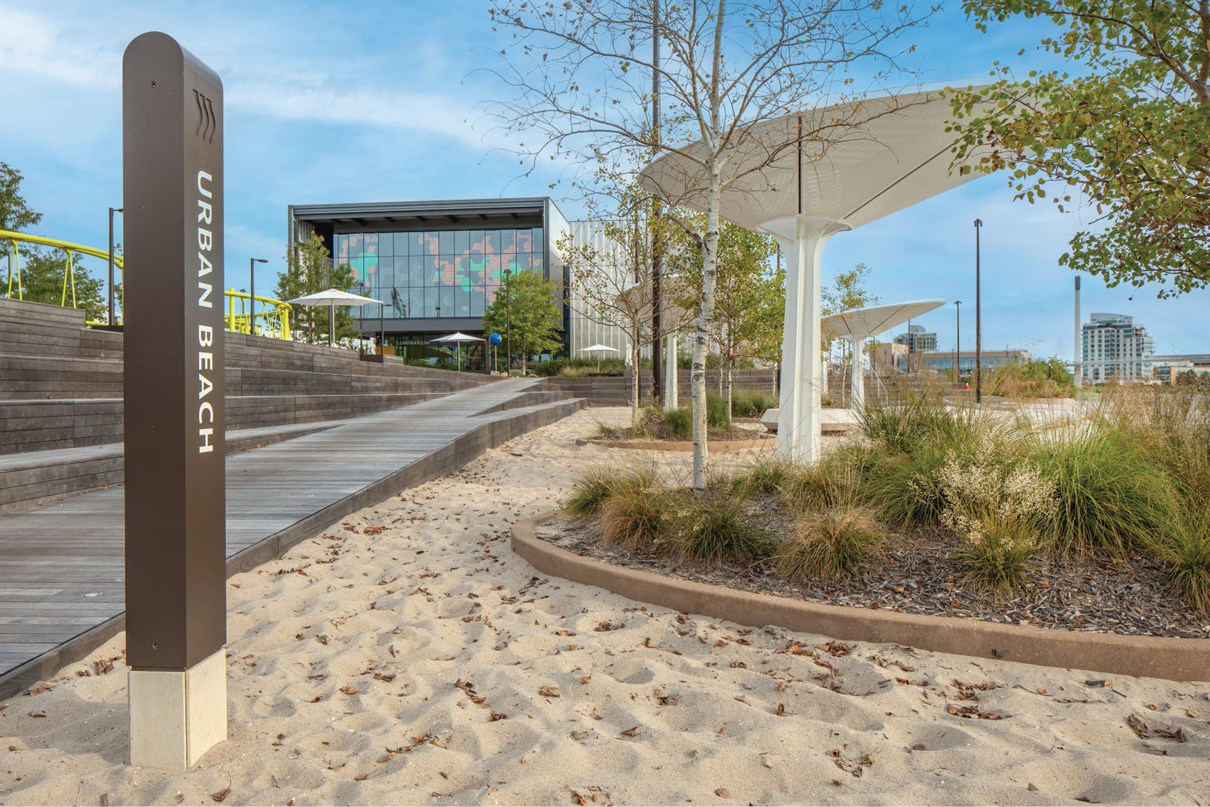 Urban Beach area marker at Lewis and Clark Landing RiverFront park in downtown Omaha, Nebraska, featuring a sandy beach, white shade canopy structures, native plantings, wood boardwalk terracing, and the Luminarium building visible in the background