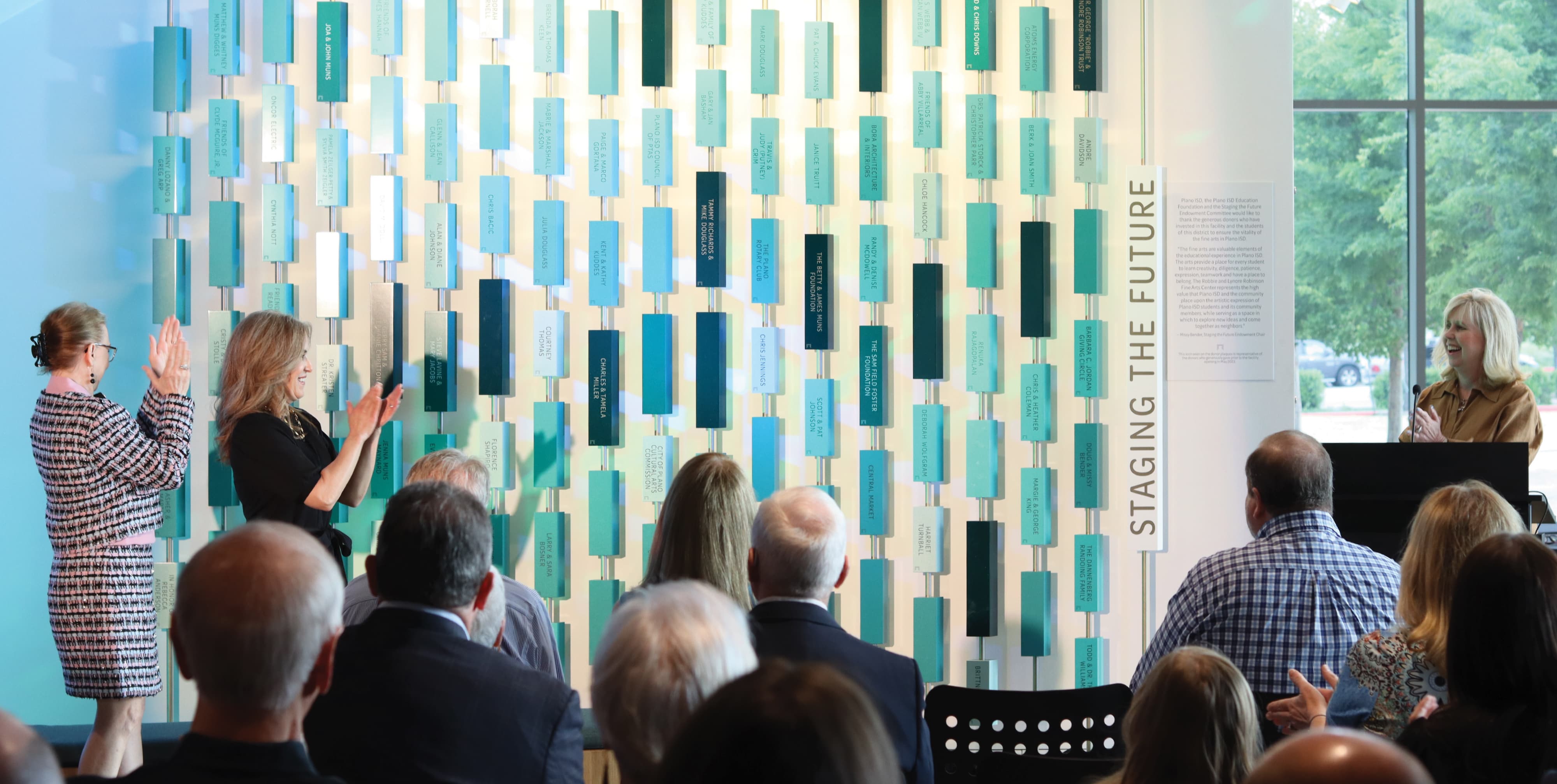 Audience gathered at a donor recognition unveiling in front of the “Staging the Future” installation, a large-scale environmental graphic design feature with suspended dimensional plaques supporting placemaking and experiential storytelling in a higher education performing arts environment.