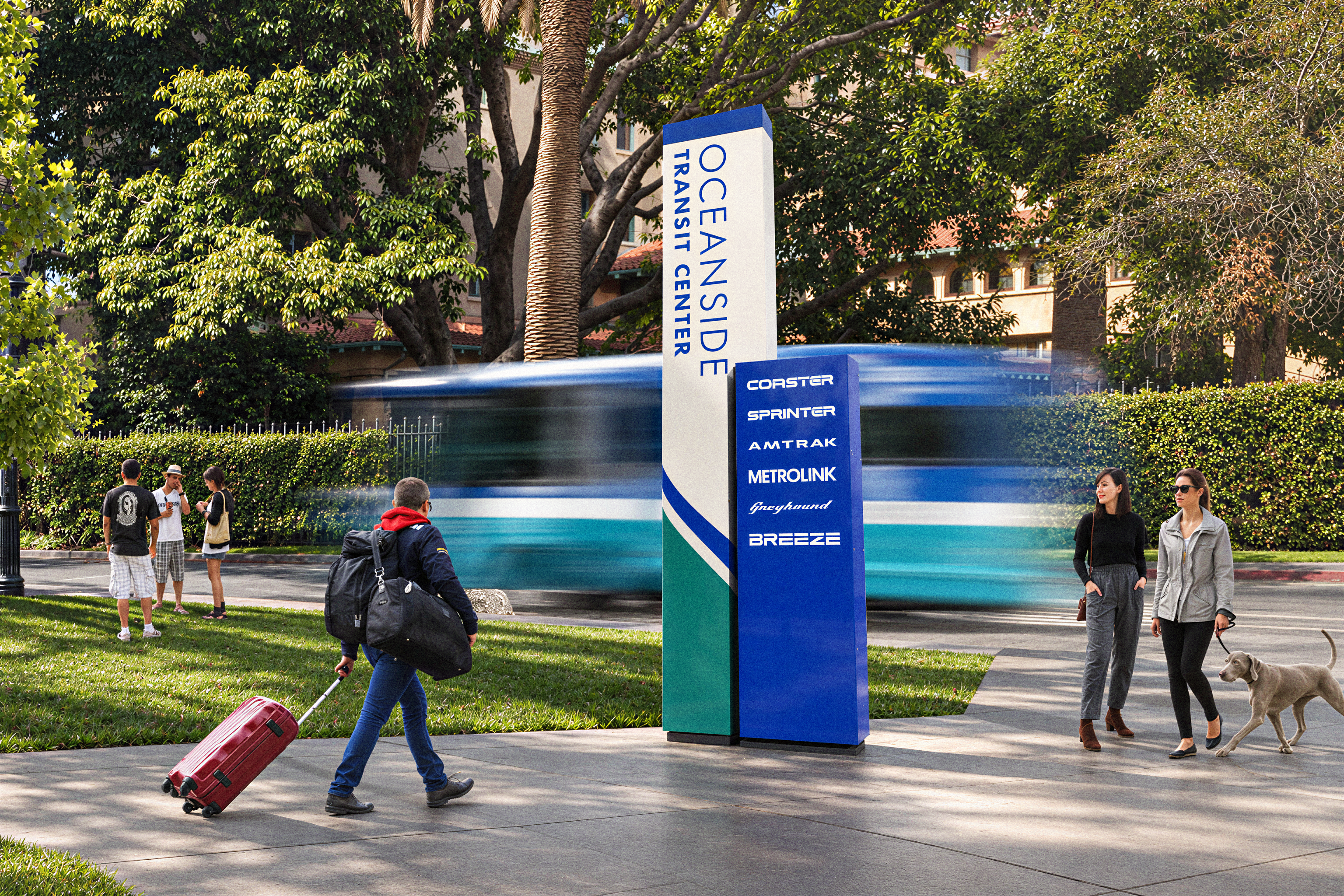Rendering of a tall blue and cream monument sign reading 'Oceanside Transit Center' with service listings for Coaster, Sprinter, Amtrak, Metrolink, Greyhound, and Breeze, as a motion-blurred train passes behind and pedestrians walk past with luggage and a dog.
