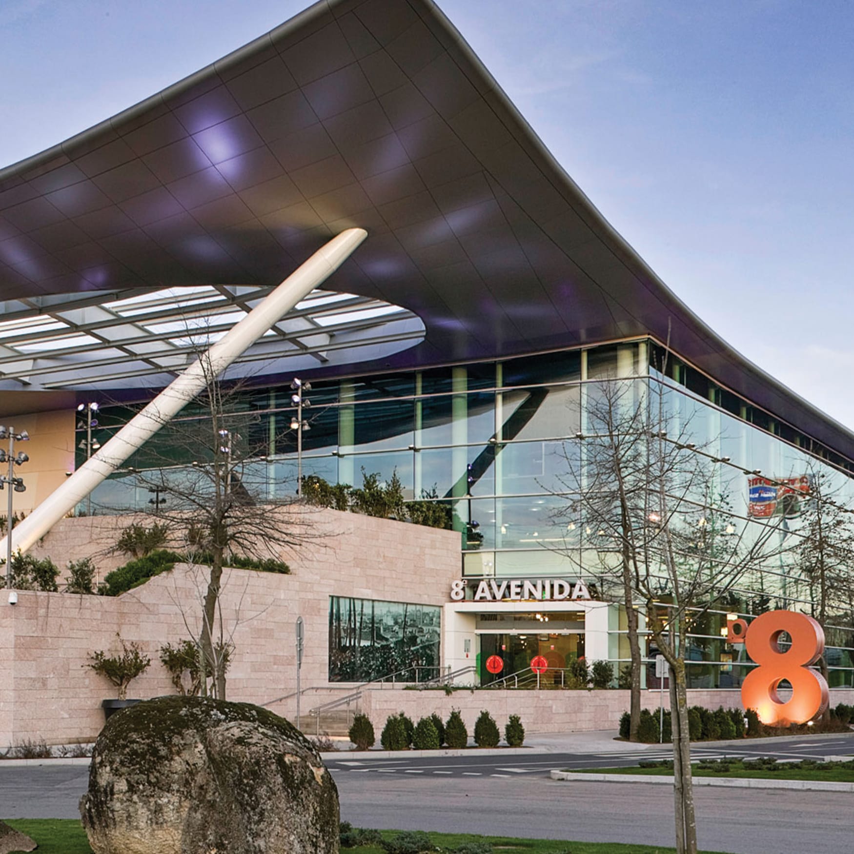 Modern exterior view of the 8 Avenida shopping center in São João da Madeira, Portugal, featuring a dramatic curved roof, glass facade, and large orange number 8 sign.