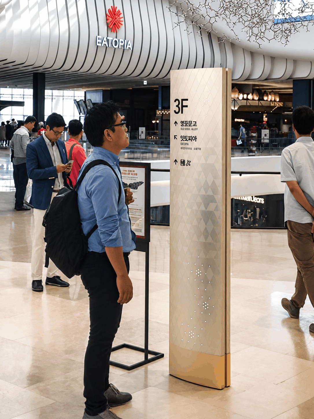 Gold '3F' directional pillar sign with Korean text and arrows, standing in a mall corridor beneath the 'Eatopia' storefront sign, with shoppers walking past storefronts and a sculptural ceiling installation overhead.