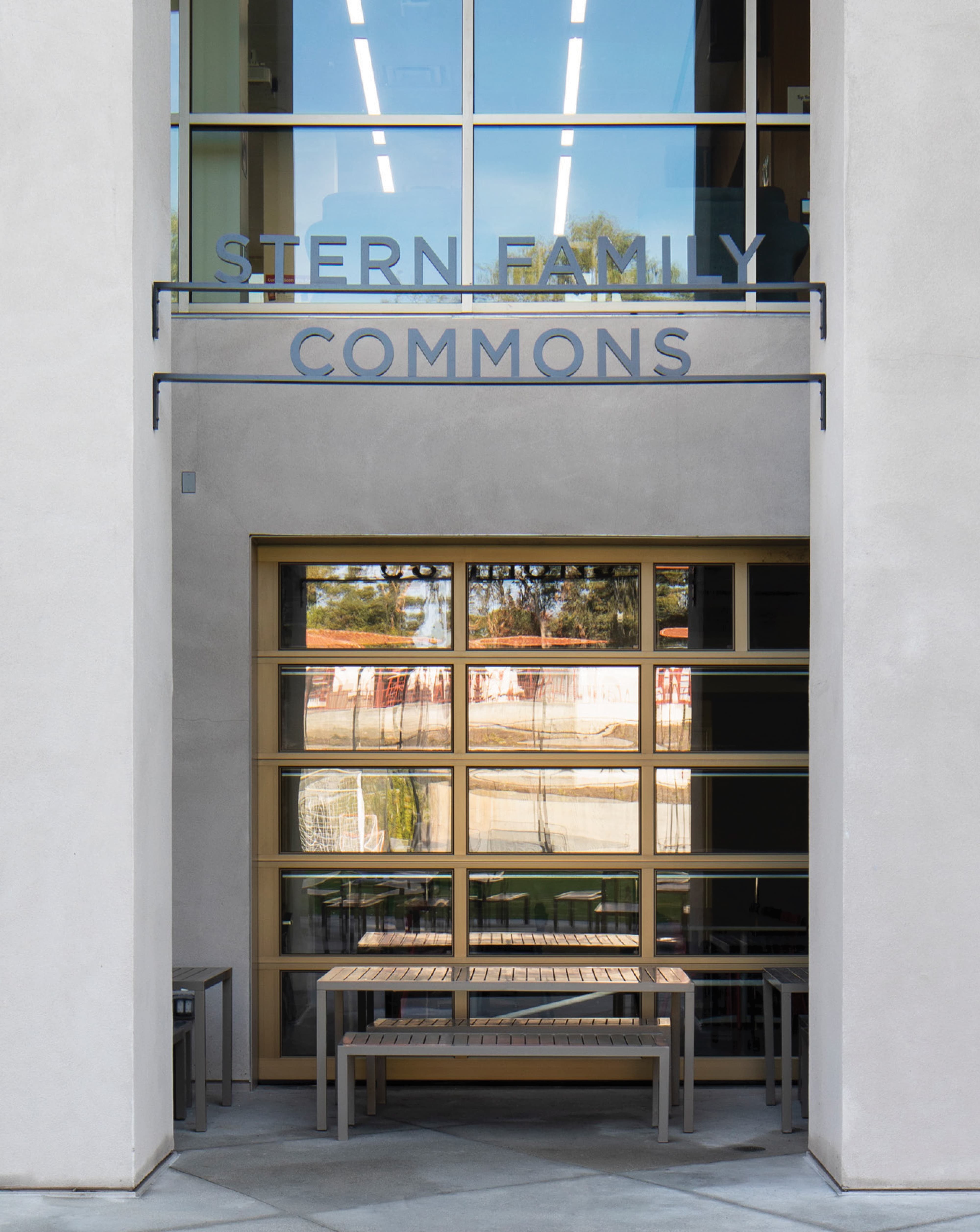 Exterior entry facade of the Stern Family Commons building featuring dimensional dark gray letters spelling the building name across two lines above a bronze-framed glass garage door, flanked by concrete columns with outdoor benches at ground level — demonstrating RSM Design's building identification signage, donor recognition and specialty graphics, and environmental graphic design services integrated seamlessly into the architecture of the education market