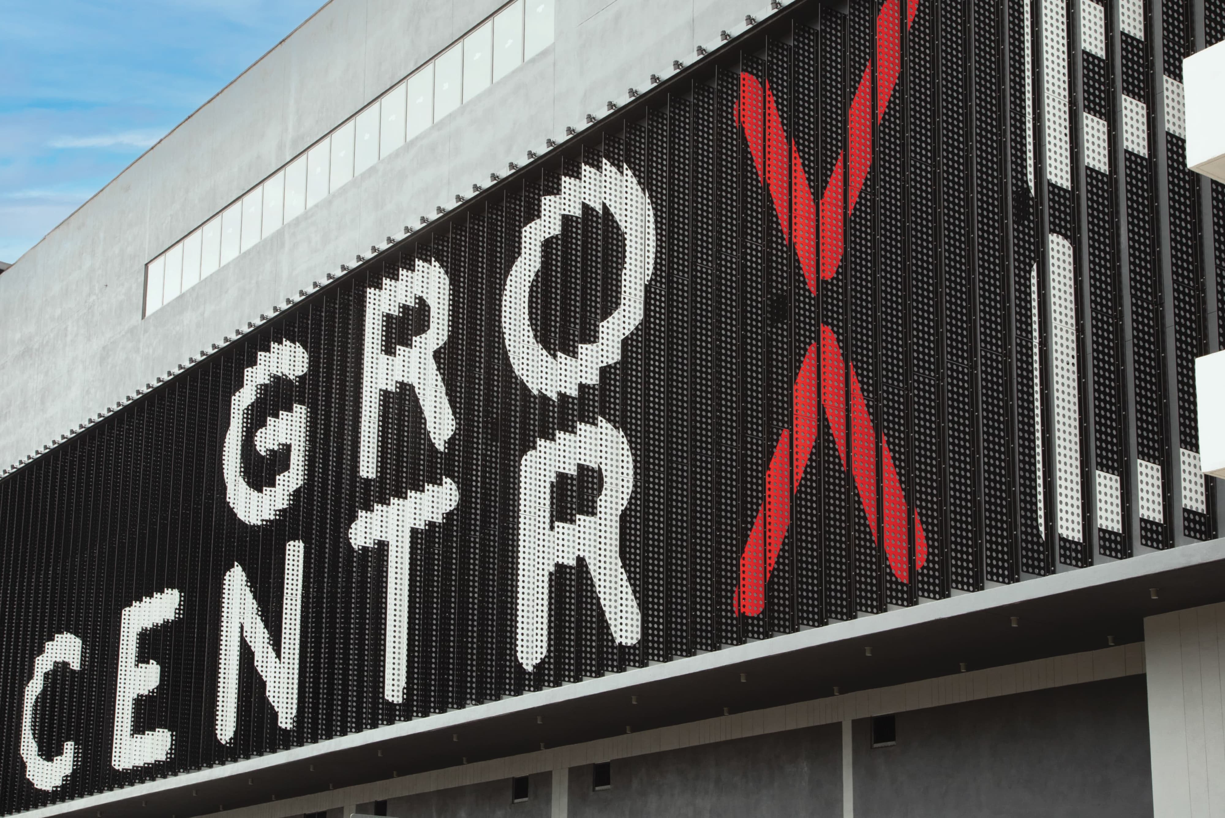 Angled close-up of the perforated metal facade at Grove Central, showing large dot-matrix letters spelling 'Grove Central' in white with a bold red X accent, set against a partly cloudy sky.