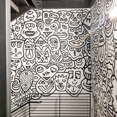 Interior of an elevator at Uncommons covered floor-to-ceiling on three walls with a bold black-and-white illustrated mural. The artwork features a dense, playful pattern of abstract characters, faces, creatures, and geometric shapes in a doodle art style.