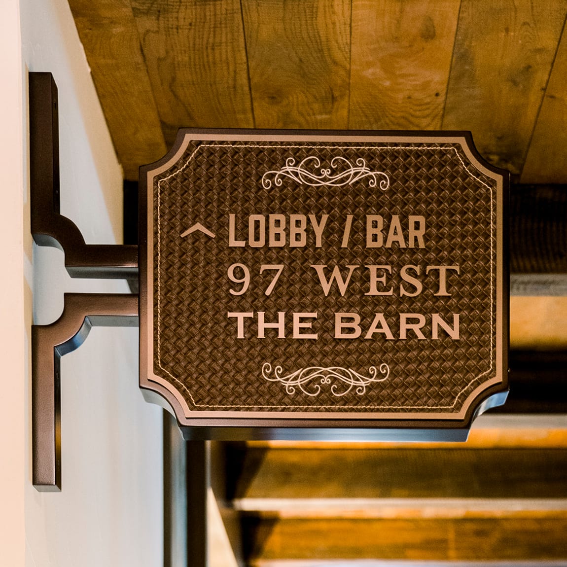 Interior wayfinding sign at Hotel Drover directing guests to the Lobby Bar, 97 West Kitchen, and The Barn event space, featuring tooled leather texture and Western-style typography with decorative scroll details