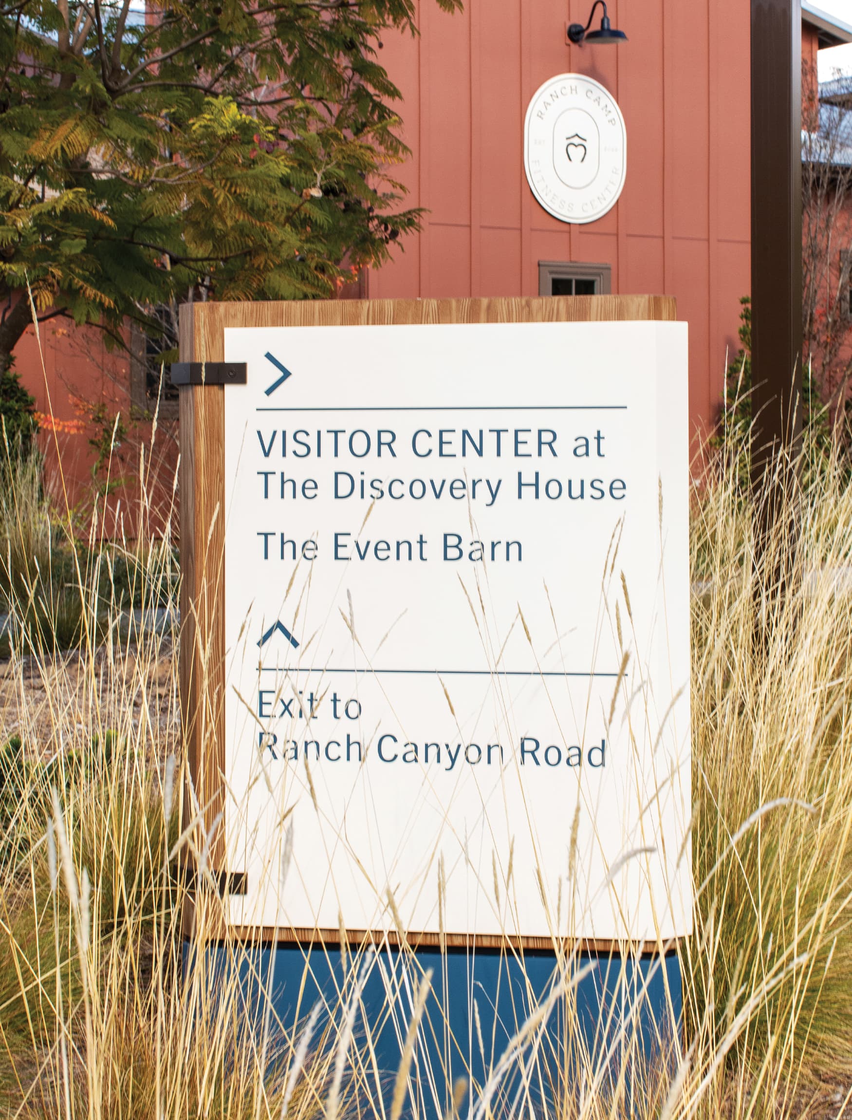 Close up detail of wooden signage with white plaque and minimal navy blue text. Ranch Camp fitness center in the background. 