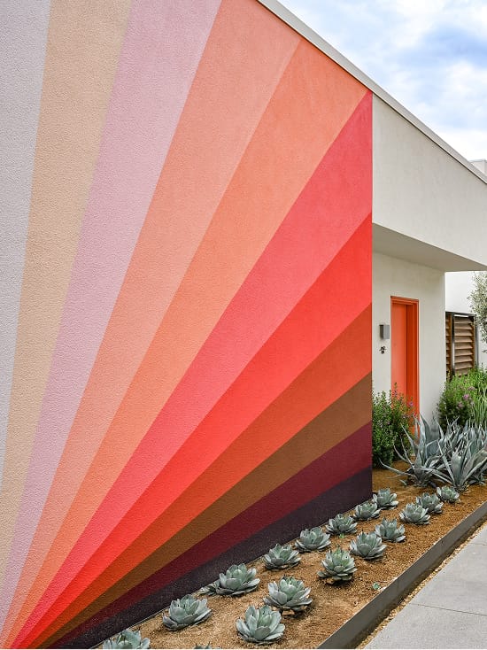 Exterior stucco wall at Thompson Palm Springs painted with a large sunburst mural in radiating stripes of pink, coral, red, and brown, with a desert planting bed of blue-grey agave and succulents and an orange guest room door at the corner.
