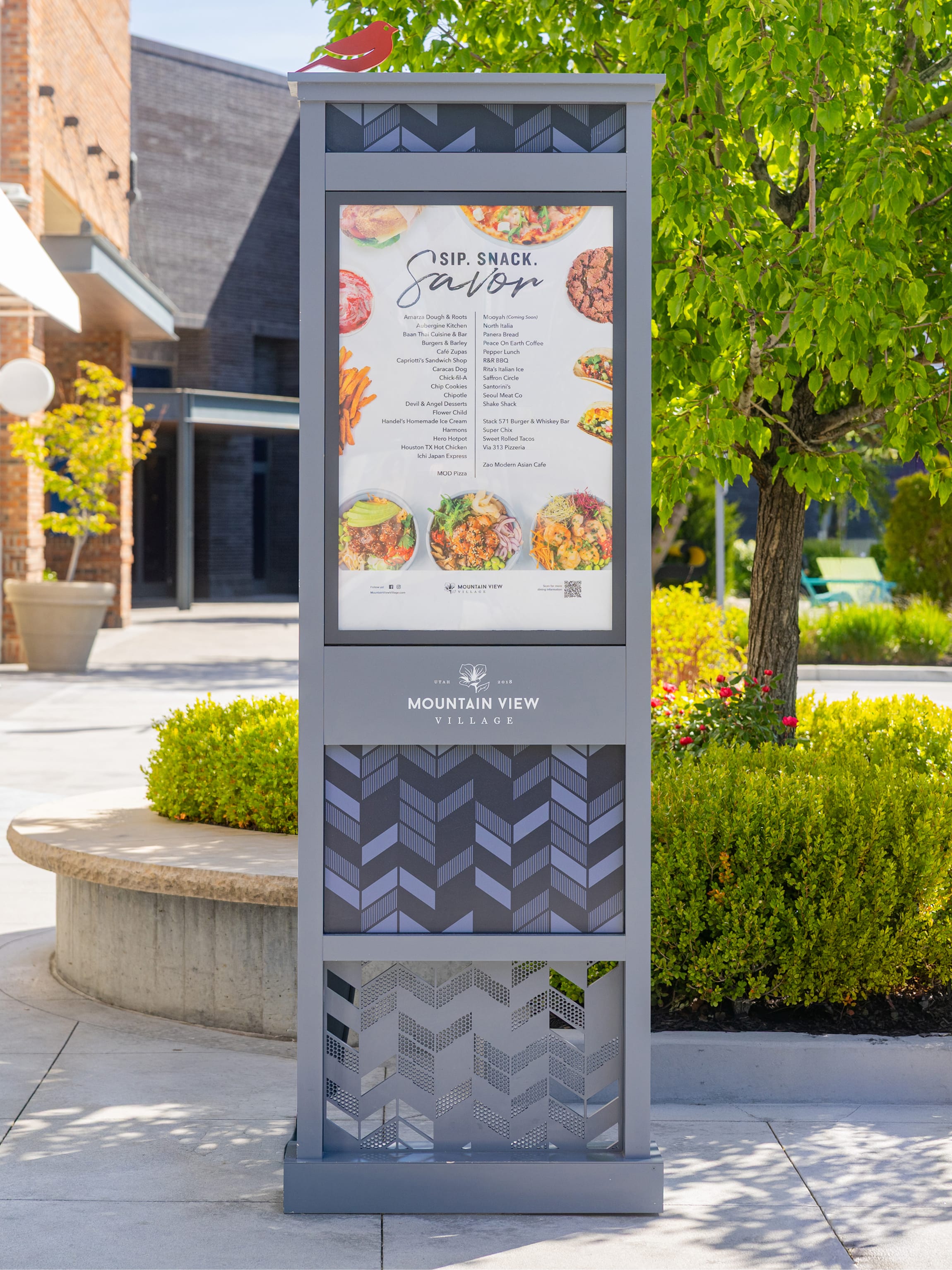 Freestanding directory kiosk with a red bird topper listing dining options under "Sip. Snack. Savor." and the Mountain View Village logo, set among landscaped seating at the Riverton, Utah center — wayfinding and environmental graphic design by RSM Design.
