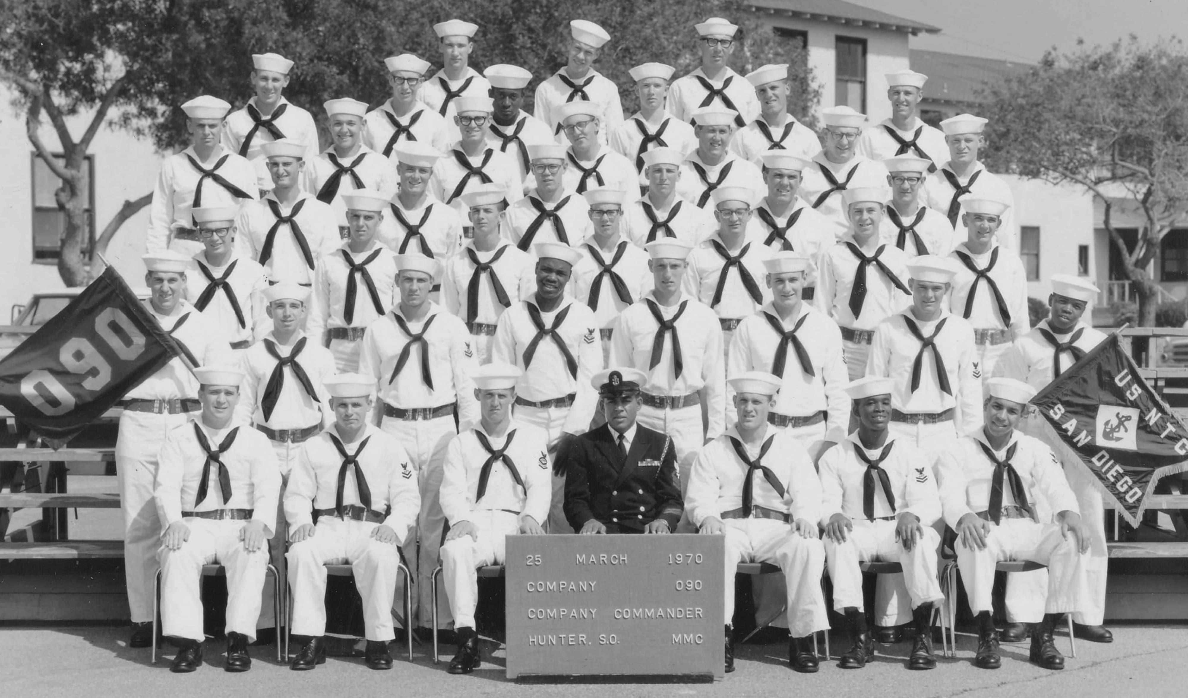 Vintage black and white group photo of Navy recruits in dress white uniforms arranged in rows, with company flags reading '090' and 'US NTC San Diego,' and a sign reading '25 March 1970, Company 090, Company Commander Hunter, S.O., MMC.