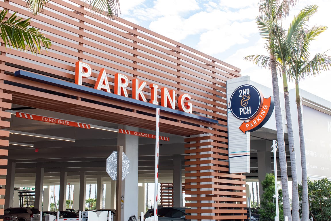 Exterior parking garage entrance at 2nd & PCH in Long Beach, California, featuring large-scale dimensional 'Parking' channel letter signage in orange mounted on a wood-slat facade, alongside a branded circular 2nd & PCH parking identification pylon sign with an orange arrow — showcasing RSM Design's wayfinding signage design, master sign program, and environmental graphic design for a coastal waterfront retail, parking garage, and mixed-use destination.