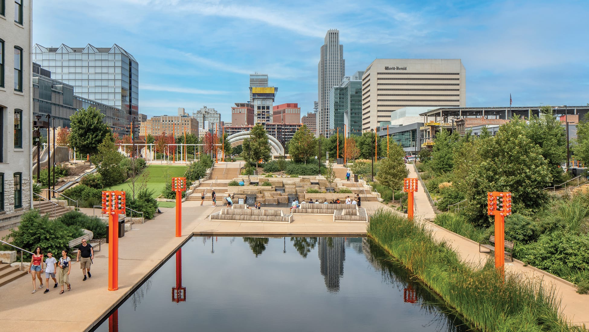 Panoramic view of the RiverFront Park and reflection pool in downtown Omaha, Nebraska, featuring orange light tower sculptures, terraced seating, lush greenery, pedestrians, and the Omaha city skyline including the World-Herald building on a sunny summer day