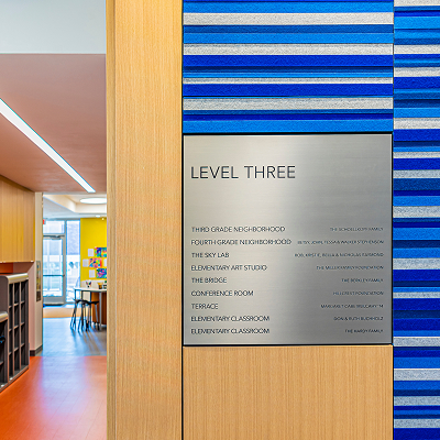 Metallic 'Level Three' directory sign listing rooms including Third Grade Neighborhood, Fourth Grade Neighborhood, The Sky Lab, Elementary Art Studio, The Bridge, Conference Room, Terrace, and Elementary Classrooms with donor names, set against a blue and silver striped wall beside a wood-paneled hallway.