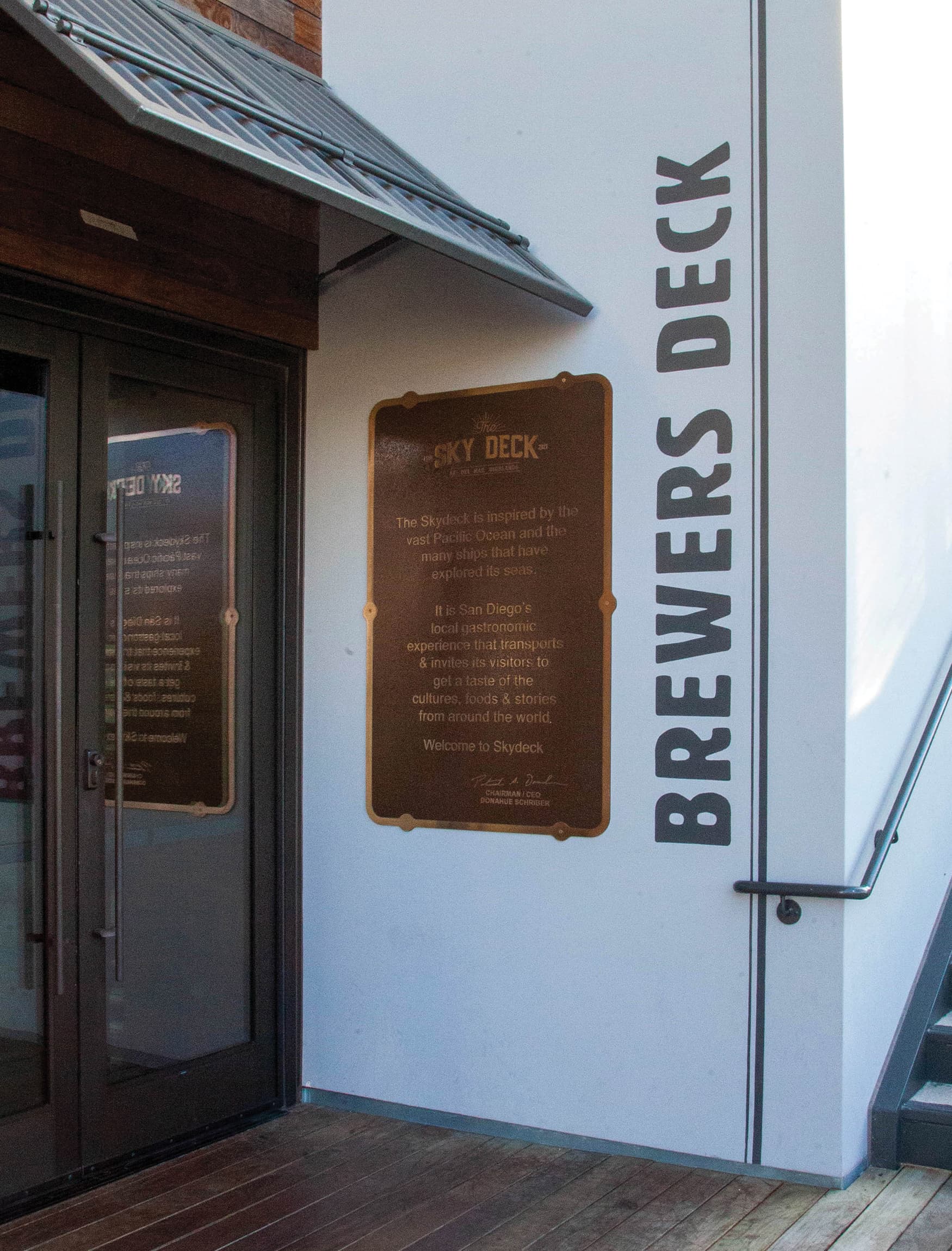 Mounted educational plaque sign and vertical painted tenant identity wall signage for The Skydeck, a retail food and beverage hall, located in Del Mar, California.
