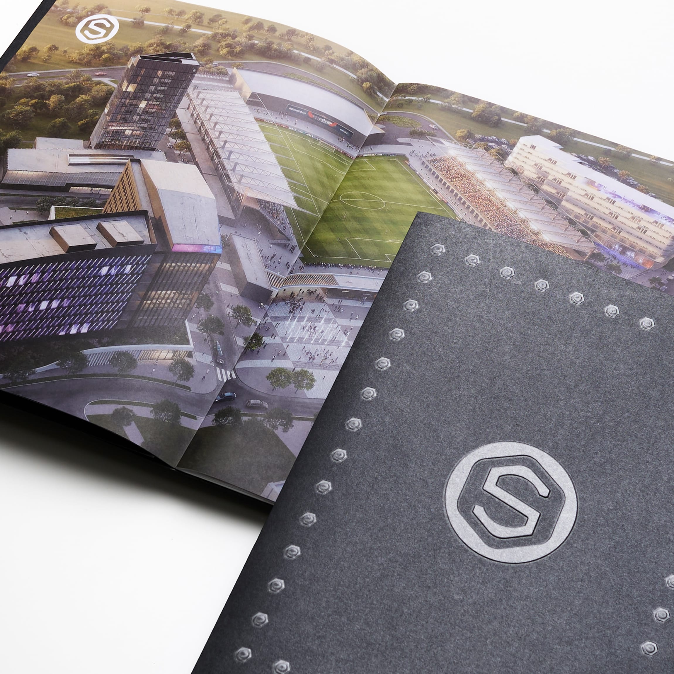 Open leasing brochure spread with an aerial rendering of a mixed-use development anchored by a soccer stadium, surrounded by office and retail buildings, layered over a charcoal cover embossed with the Staybolt Street mark and bolt motif, Staybolt Street, Mansfield, Texas, brand identity and leasing collateral design by RSM Design