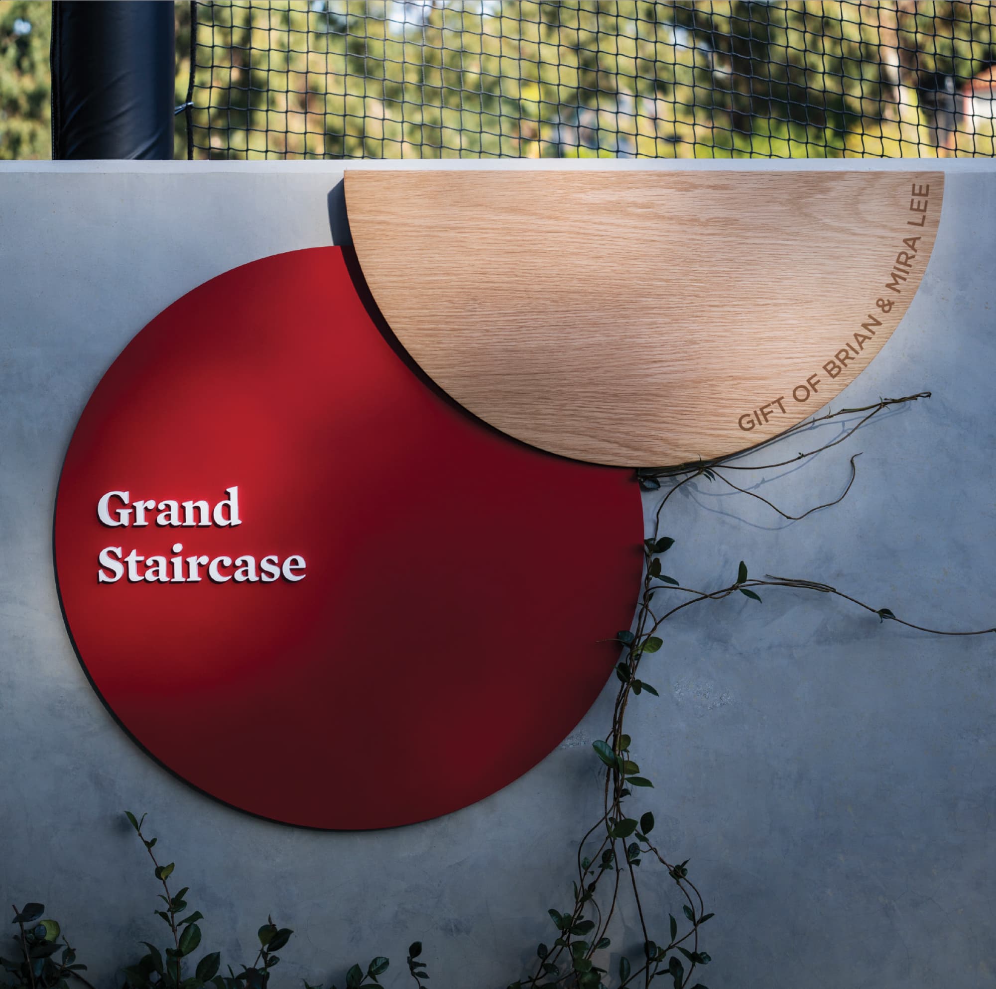 A sculptural donor recognition and space identification sign for the "Grand Staircase" composed of an overlapping red metal disc with dimensional white lettering and a natural oak wood half-circle engraved with "Gift of Brian & Mira Lee," mounted on a concrete wall with climbing vines — exemplifying RSM Design's donor recognition and specialty graphics services that balance wayfinding and philanthropic acknowledgment with artistic material expression within the education market