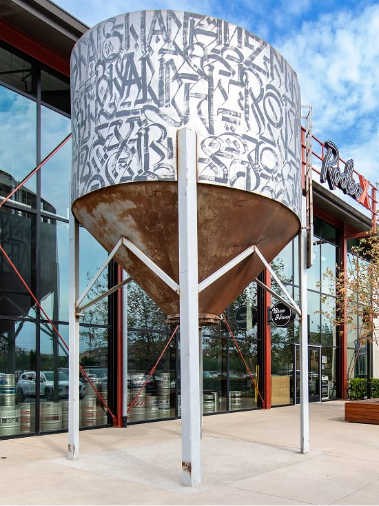 Close-up of the repurposed grain silo painted with abstract calligraphic graffiti lettering, mounted on white support beams outside the glass-walled 'Rodeo X' building, with the 'Brew Haven' sign visible through the windows.
