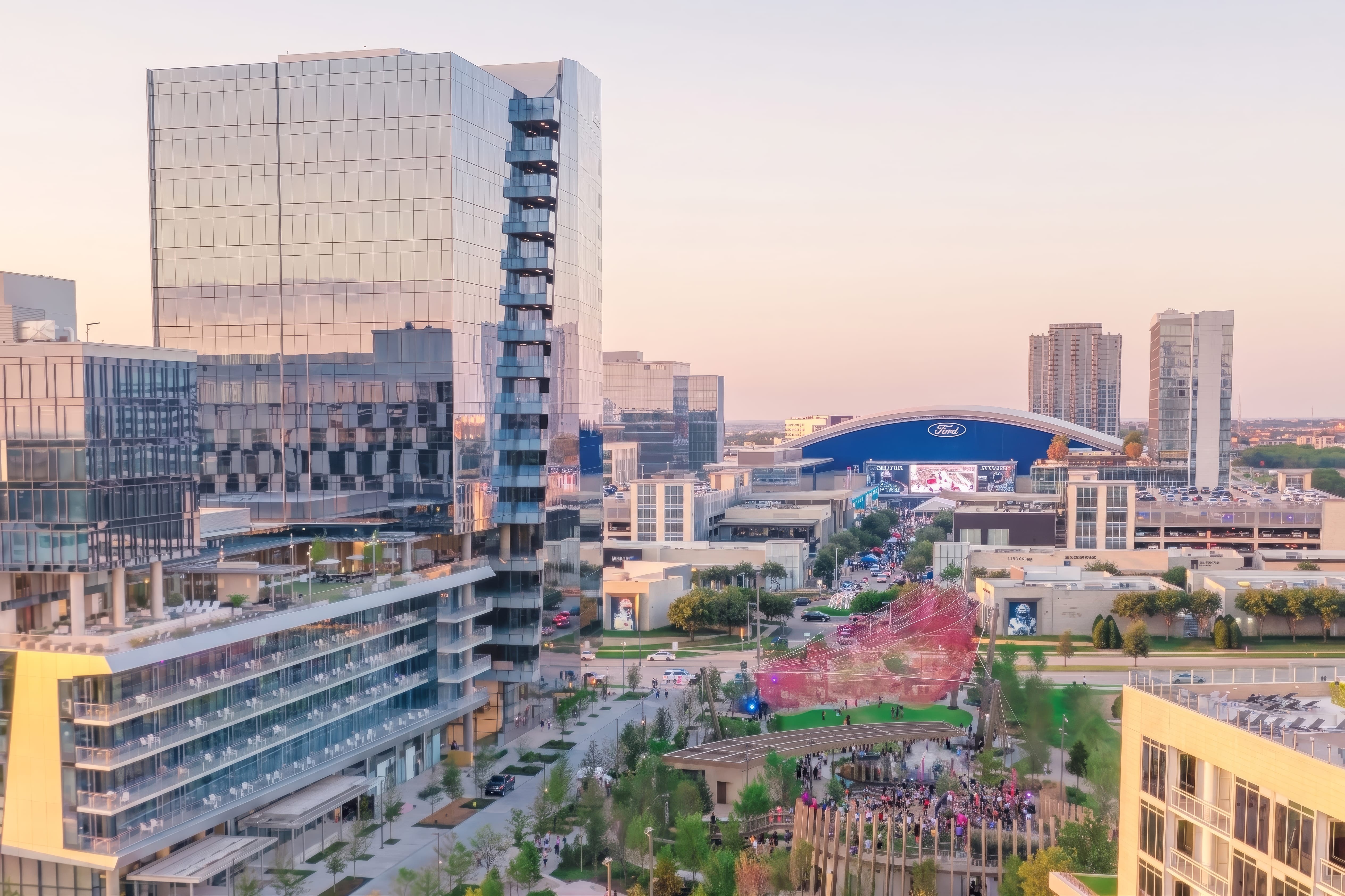 Aerial dusk view of Kaleidoscope Park mixed-use urban development in Dallas, Texas with glass office towers, outdoor event lawn with pink art installation, pedestrian plaza, and Ford Center arena in background