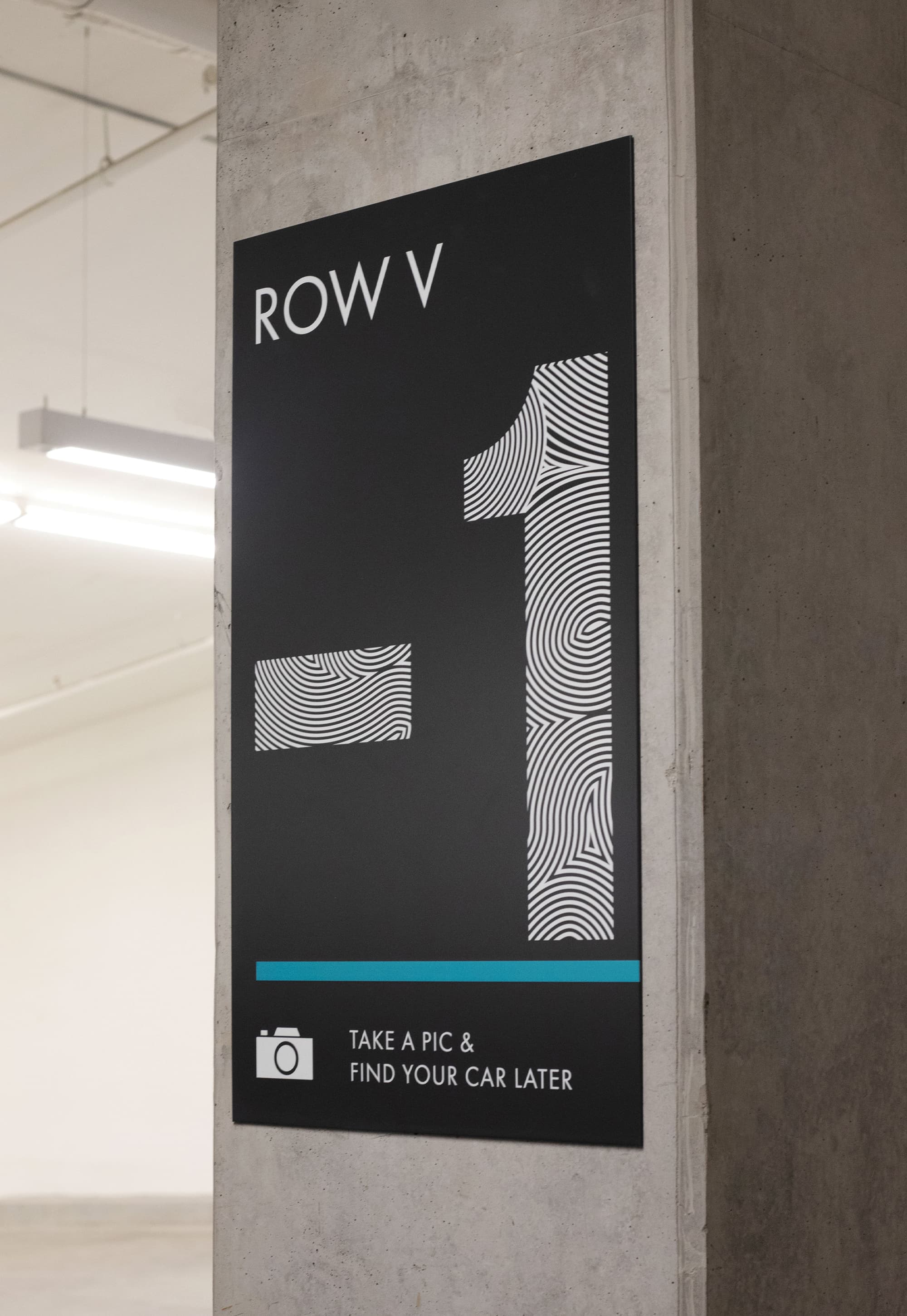 Black parking garage column sign reading 'Row V, -1' with fingerprint-patterned numerals, a blue accent stripe, and a 'Take a pic & find your car later' reminder with a camera icon, mounted on a concrete pillar.