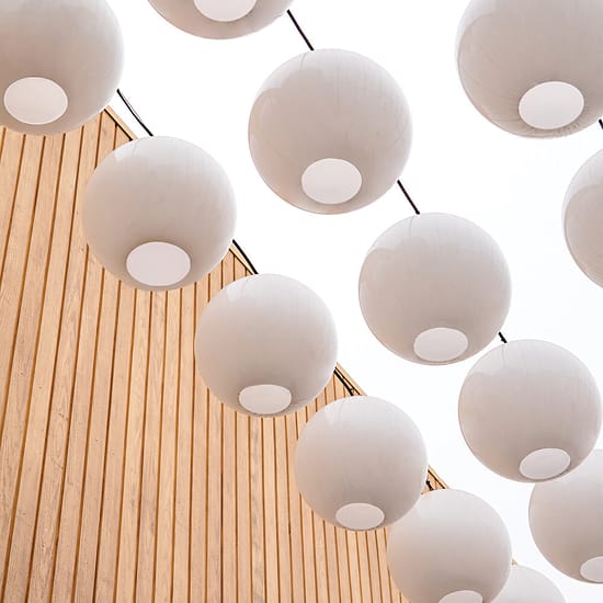 Close-up upward view of oversized white globe pendant lights suspended overhead against a bright sky, with warm wood slat cladding visible along the building facade at Rodeo 39 Public Market in Stanton, CA.