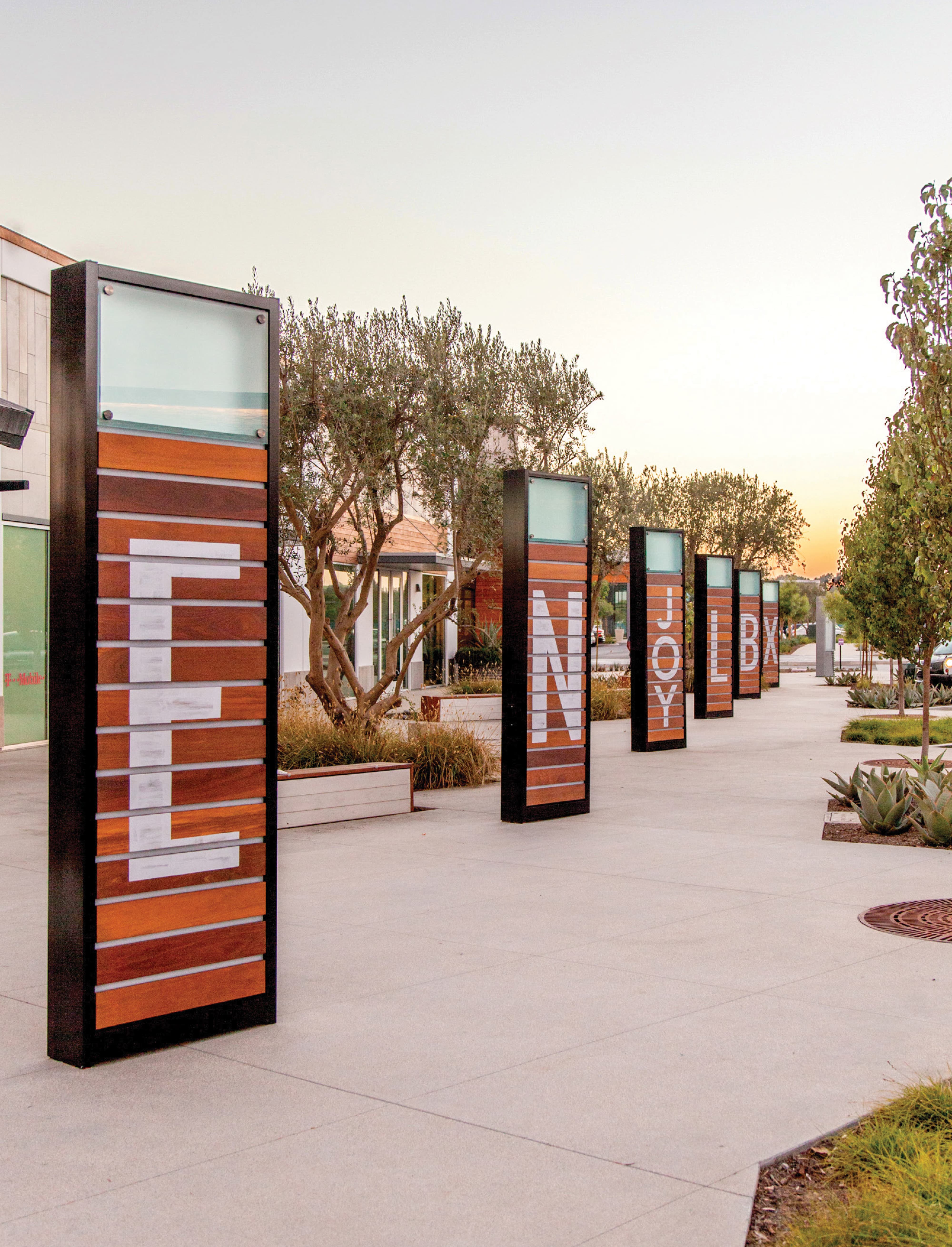 Row of tall wood-slat pylon signs receding down a paved plaza, spelling out words like 'Feel' and 'Joy' in white letters, with 'LBX' visible on a farther pylon, framed by olive trees and agave landscaping at dusk.