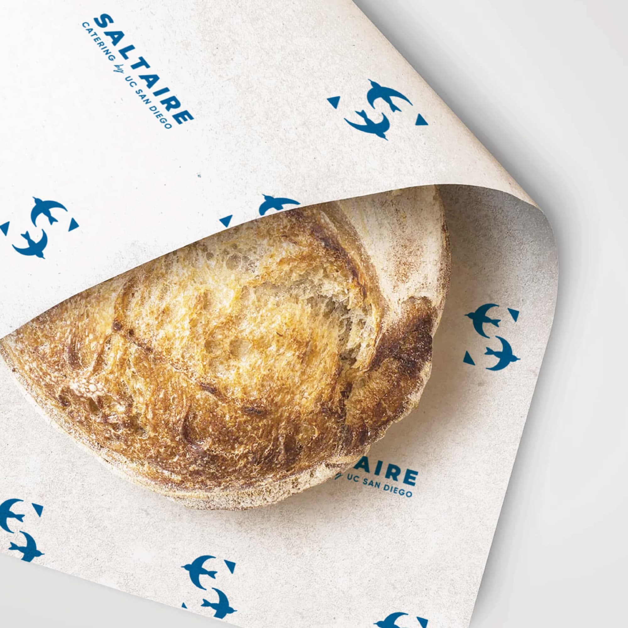 Artisan sourdough loaf wrapped in Saltaire Catering by UC San Diego branded tissue paper featuring a repeating tern lettermark pattern in navy, showcasing specialty graphics and print collateral design for a food and beverage and education market client by RSM Design.