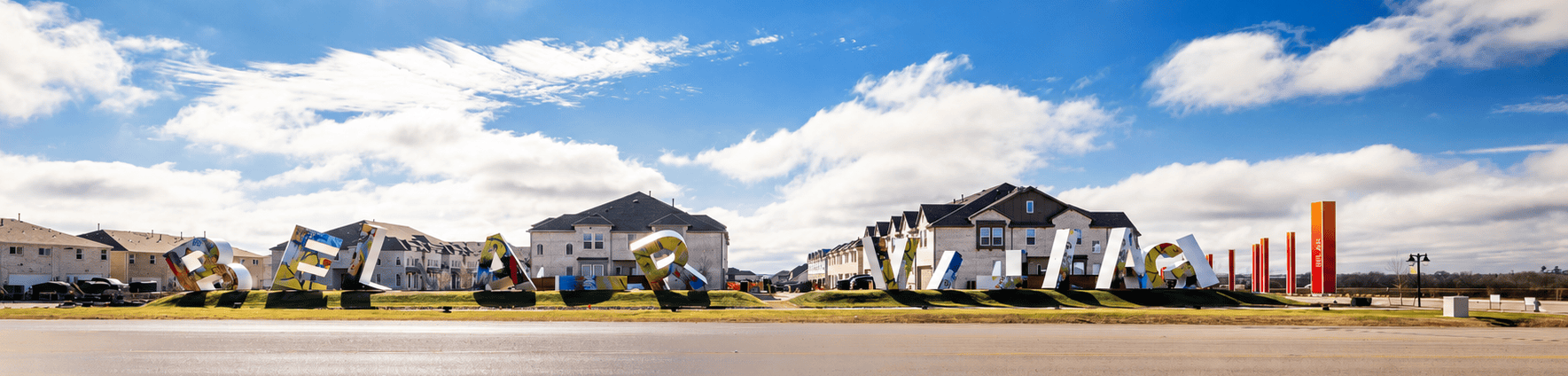 Taken from the street facing a suburban neighborhood of large, brick, 2.5-story, multi-family homes. The letter sculptures stand on a grassy strip between the street and the neighborhood. The illuminated pylons wrap around the corner of the block.