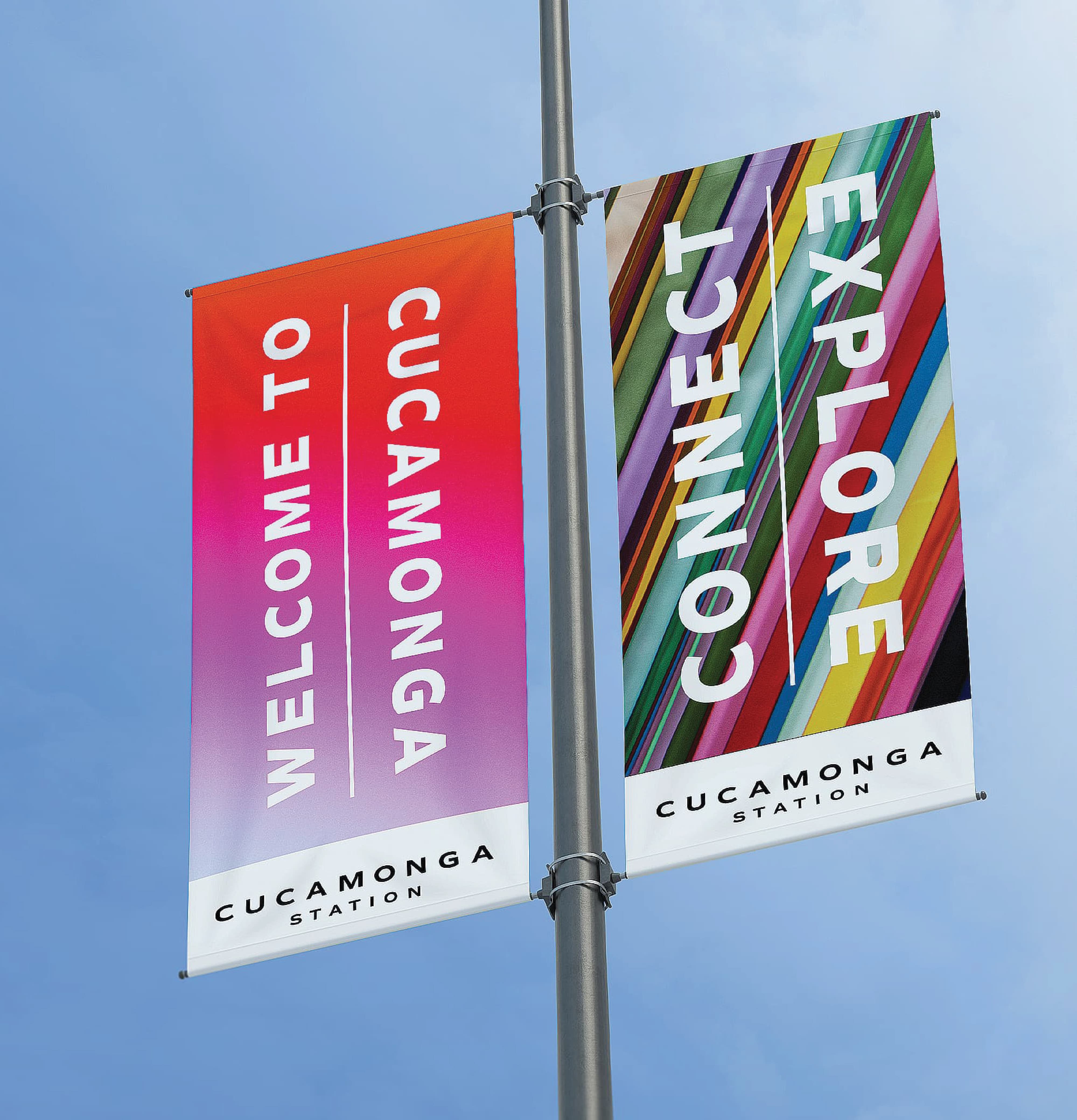 Two double-sided Cucamonga Station branded light pole banners displayed against a blue sky — one featuring a warm red-to-magenta gradient with vertical "Welcome to Cucamonga" typography, and the other displaying bold "Connect Explore" text over a multicolored diagonal stripe pattern — demonstrating RSM Design's branding and visual identity, experiential graphics and branded environments, and print collateral services applied to placemaking within the transit and streetscape markets
