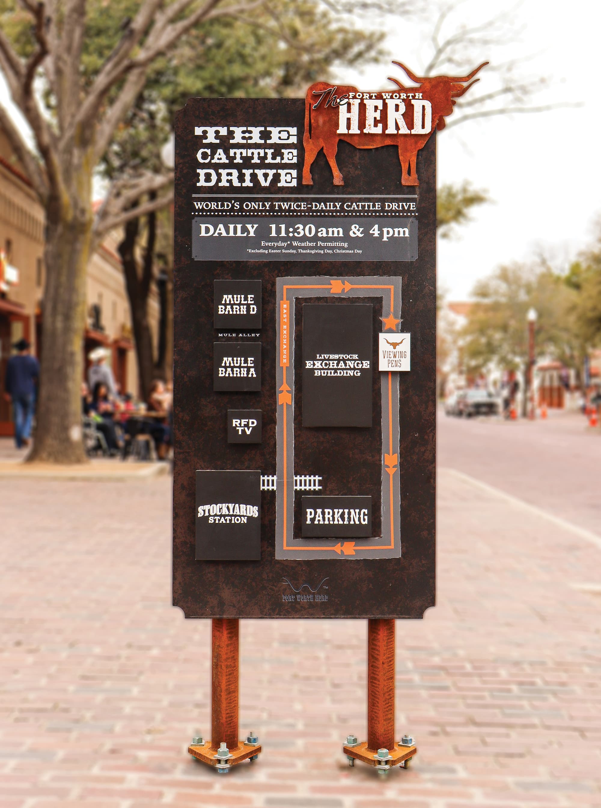 A free-standing sign made of weathered, rust-brown sheet metal, anchored into the brick sidewalk with two thick, weathered industrial pipes and pipe fittings. Text in all capital letters on the front of the sign reads, “The Fort Worth Herd. The Cattle Drive. World’s only twice-daily cattle drive. Daily 11:30am & 4pm. Everyday* Weather Permitting. *Excluding Easter Sunday, Thanksgiving Day, Christmas Day.” A crude map below the text shows the locations of “Mule Barn D”, “Mule Barn A”, “RFD TV”, “Stockyards Station”, “Livestock Exchange Building”, “Viewing Pens”, and “Parking.”
