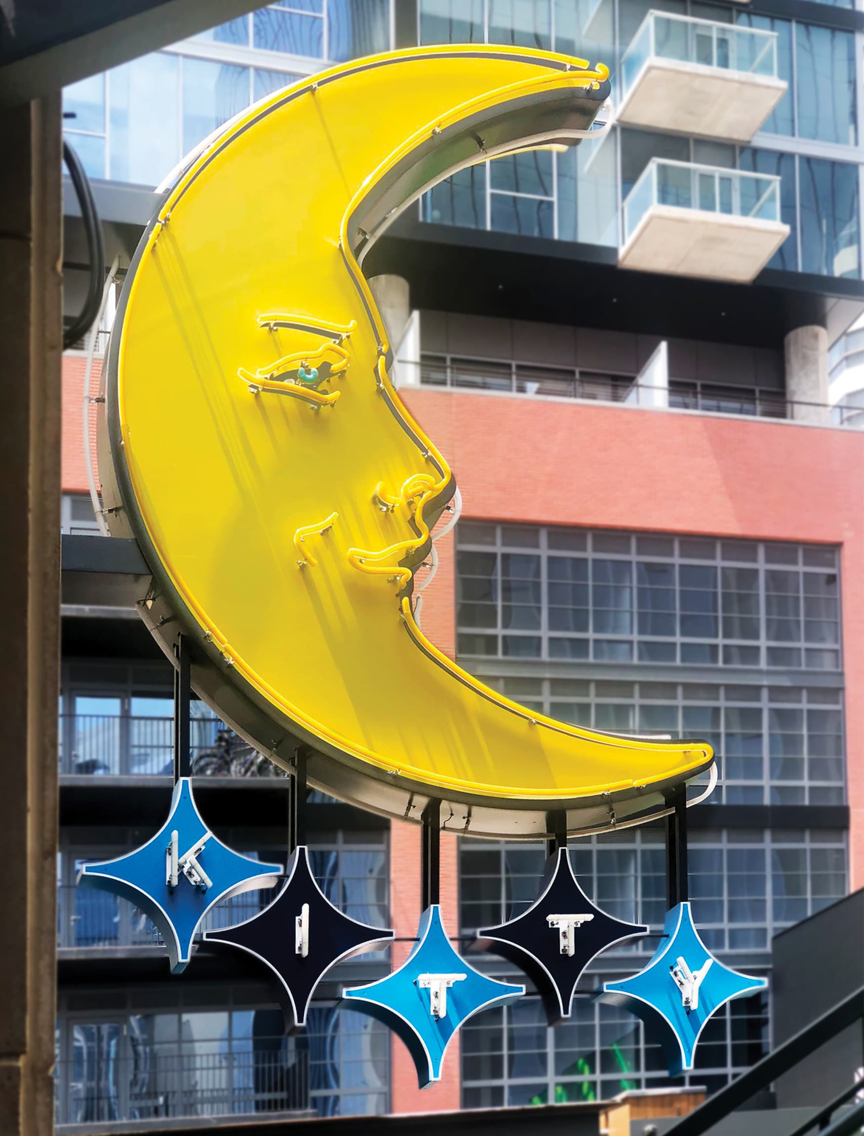Closeup of Kitty Moon memorial blade sign. Yellow crescent moon with neon tubing outlining the moon and creating a face with alternating blue and black stars hanging from bottom of moon with KITTY spelled out on the stars. Neon tube lights making up the letters of "KITTY". Signage and wayfinding for Fifth + Broadway in Nashville, Tennessee by RSM Design 