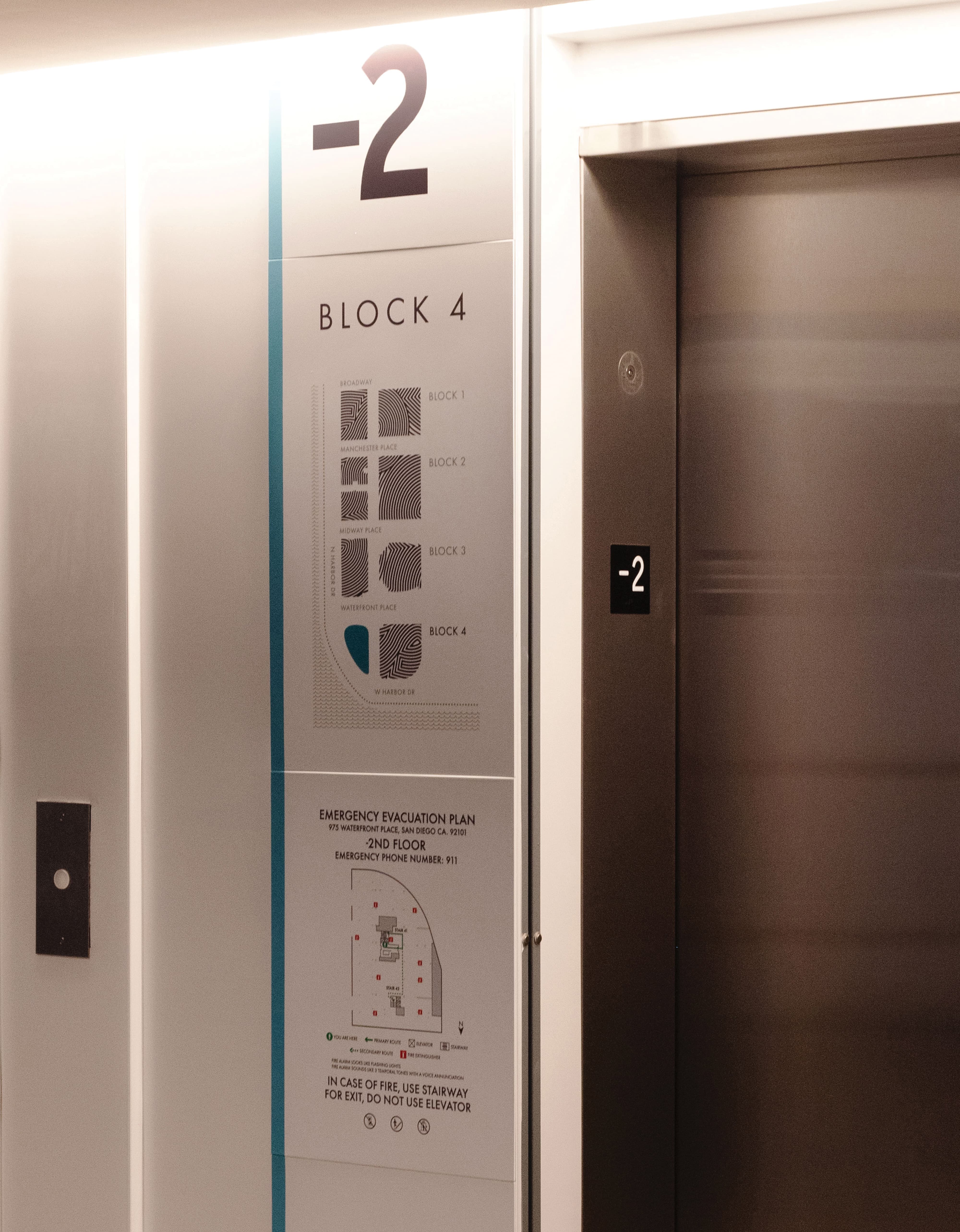 Elevator lobby signage reading '-2, Block 4,' with a fingerprint-style block map showing Blocks 1 through 4 along Broadway, Manchester Place, Midway Place, and Waterfront Place, and an emergency evacuation floor plan below, beside brushed metal elevator doors marked '-2.