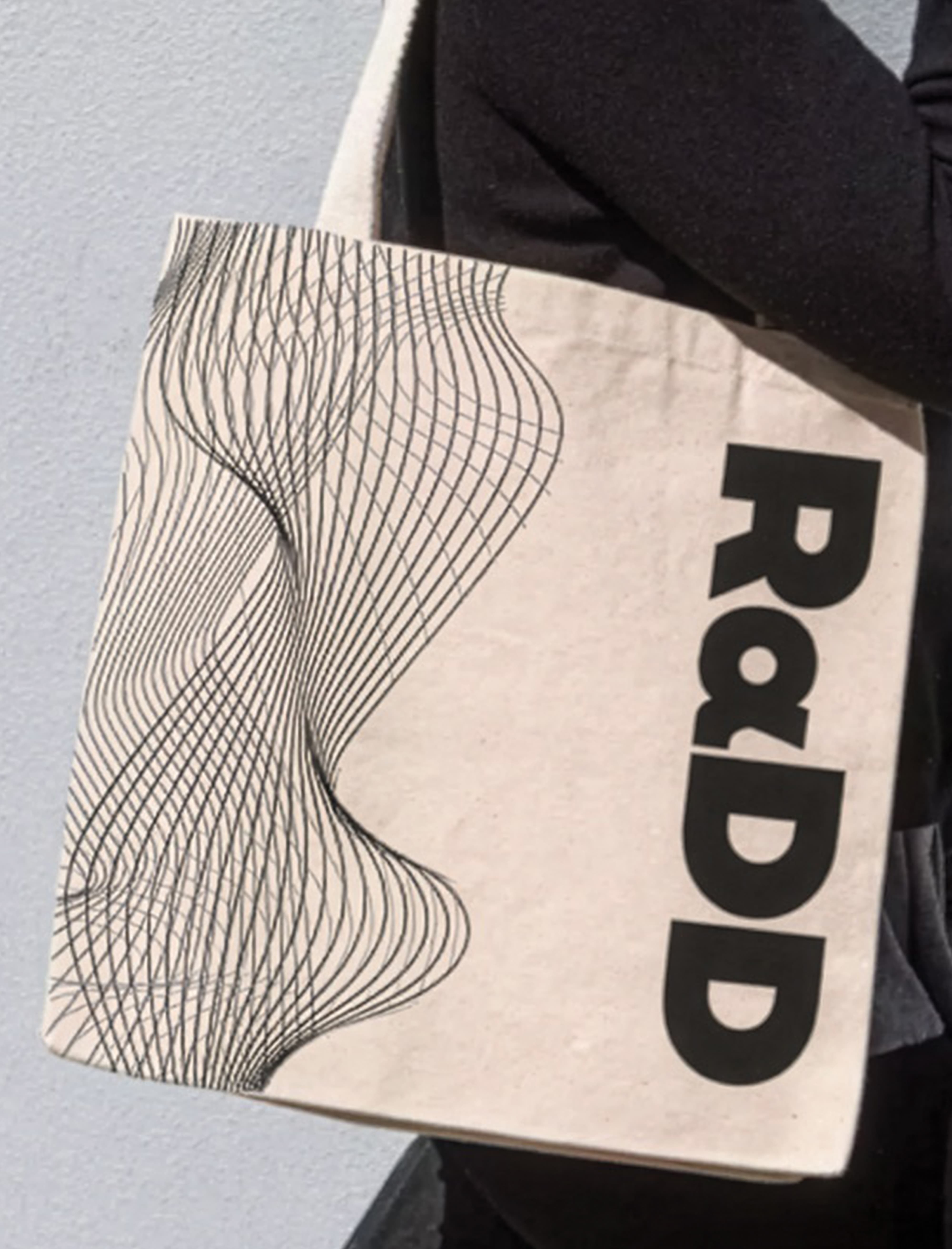 Close-up of a canvas tote bag with a black wavy line-art pattern on one side and the 'RαDD' wordmark printed vertically on the other, slung over a person's shoulder against a gray wall.