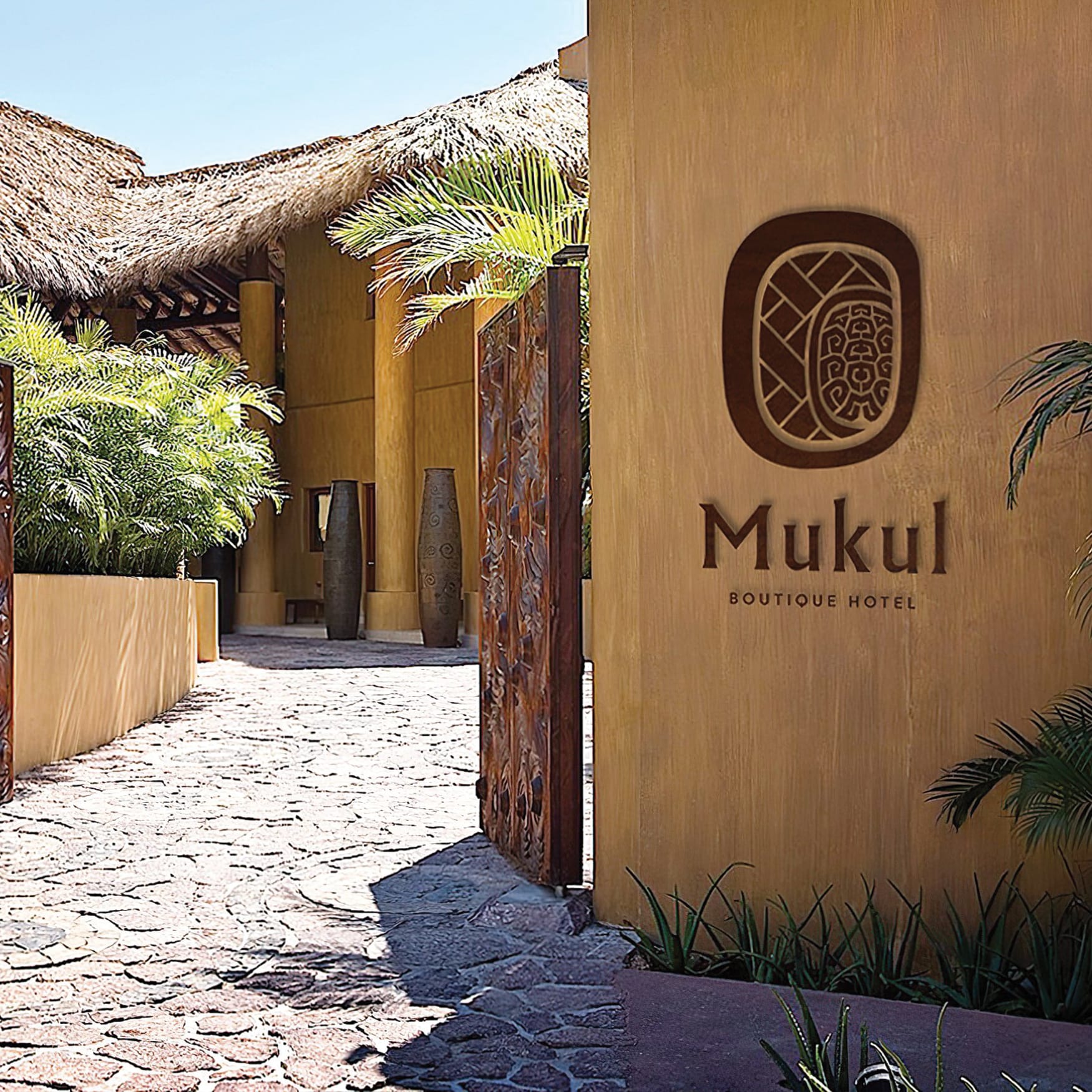 Luxury tropical boutique hotel entrance featuring a carved wooden gate, warm stucco walls, lush palm landscaping, stone pathway, and a wall-mounted “Mukul Boutique Hotel” logo sign in a serene resort setting.