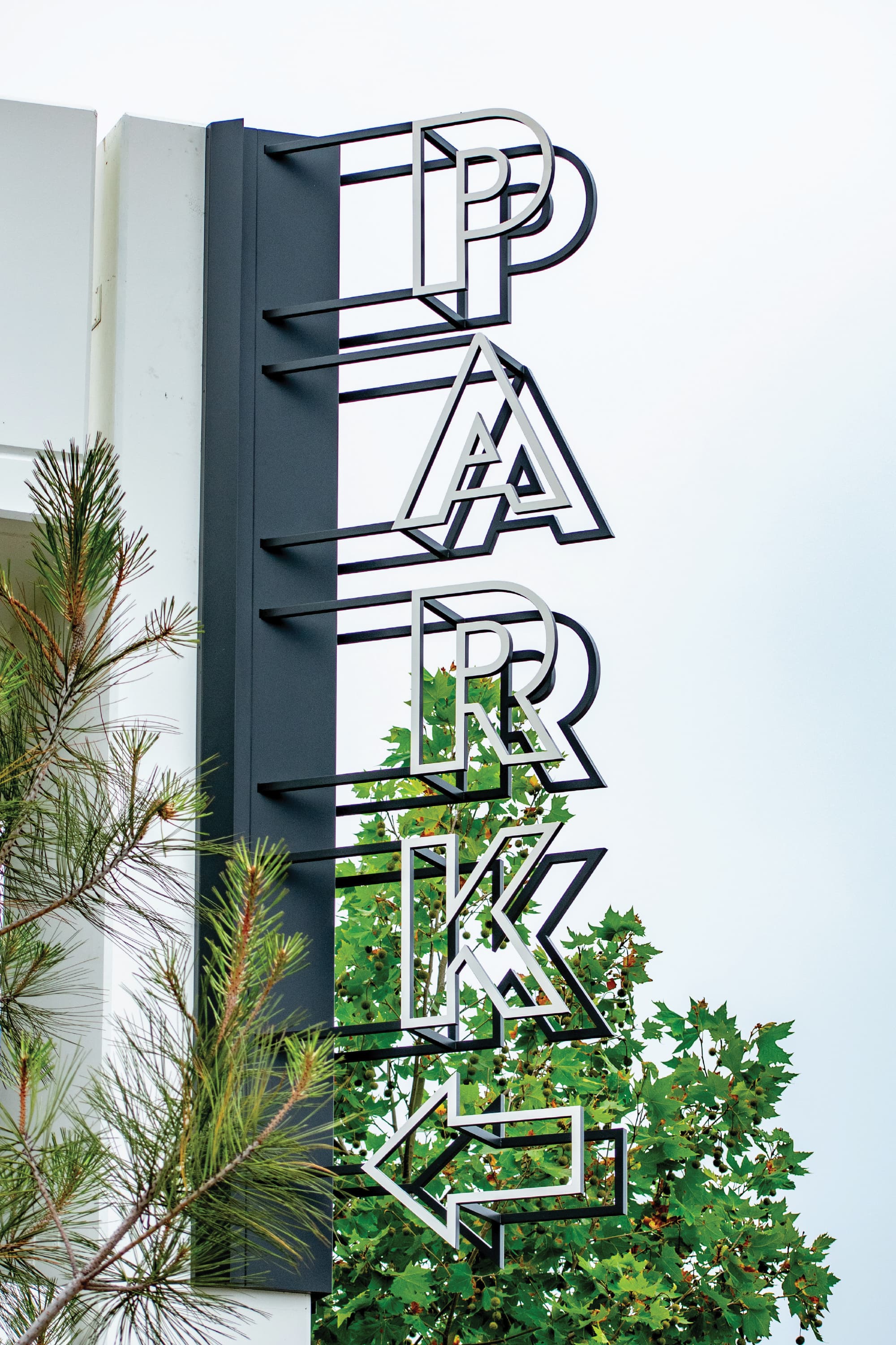 Close-up detail of a large-scale vertical dimensional "Parking" blade sign with decorative striped letterforms mounted on a dark charcoal fin panel at Dana Point Harbor, surrounded by coastal pine and oak trees — RSM Design wayfinding and signage design, master sign programs, specialty graphics, and environmental graphic design for waterfront, parking garage, civic, and mixed-use destinations.